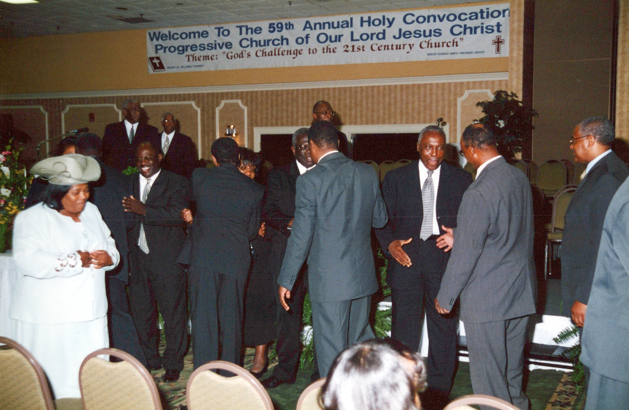A group of African American men in suits and a woman in a white suit and hat socialize at a church event under a large welcome banner.