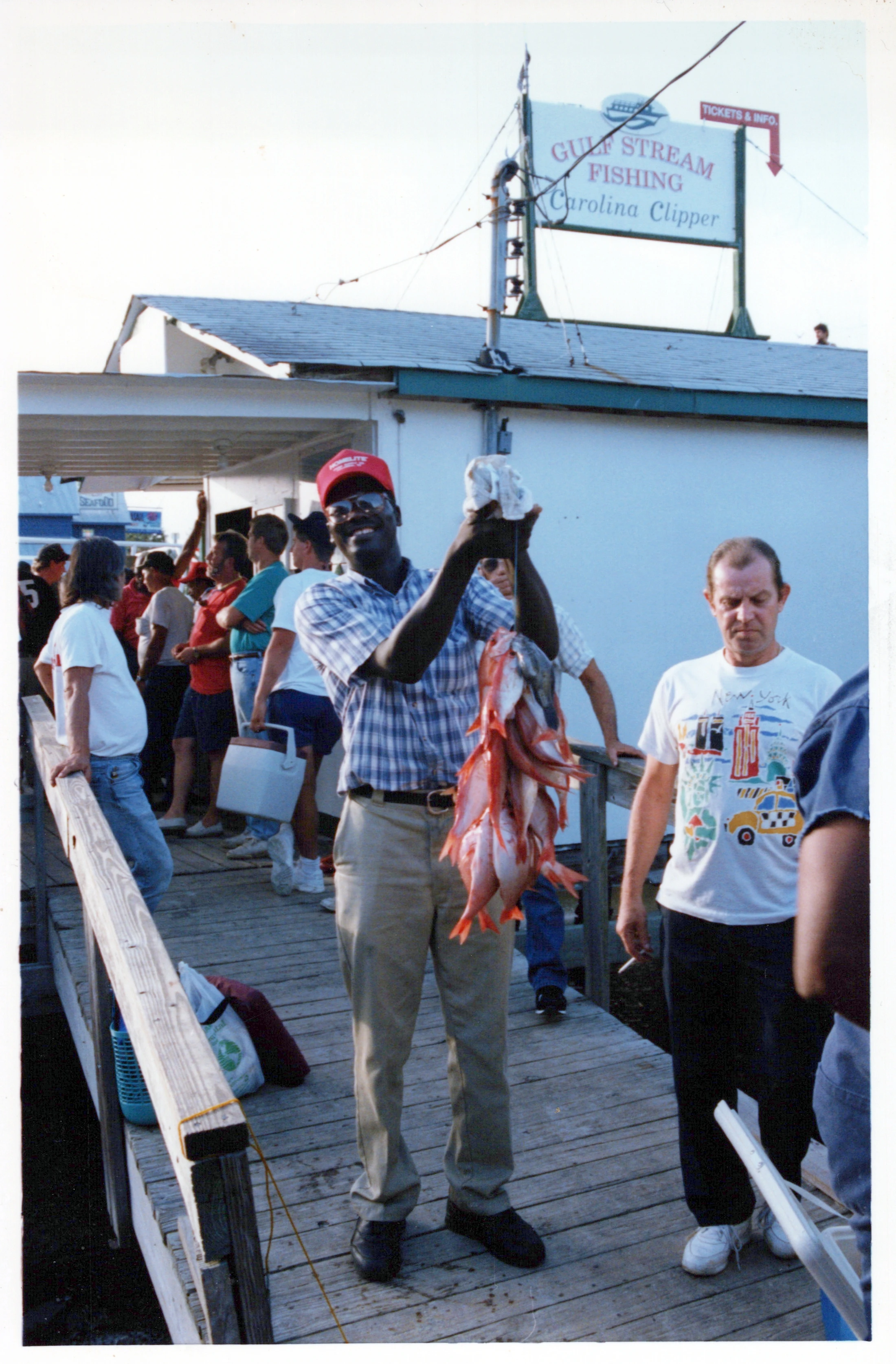 A smiling man in a red cap proudly holds up a large string of red fish. He stands on a wooden dock with other people near a fishing charter building.