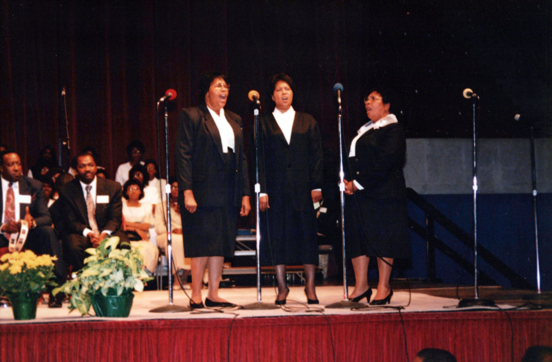 A vintage photograph of three Black women in matching dark suits singing into microphones on a stage in front of an audience.