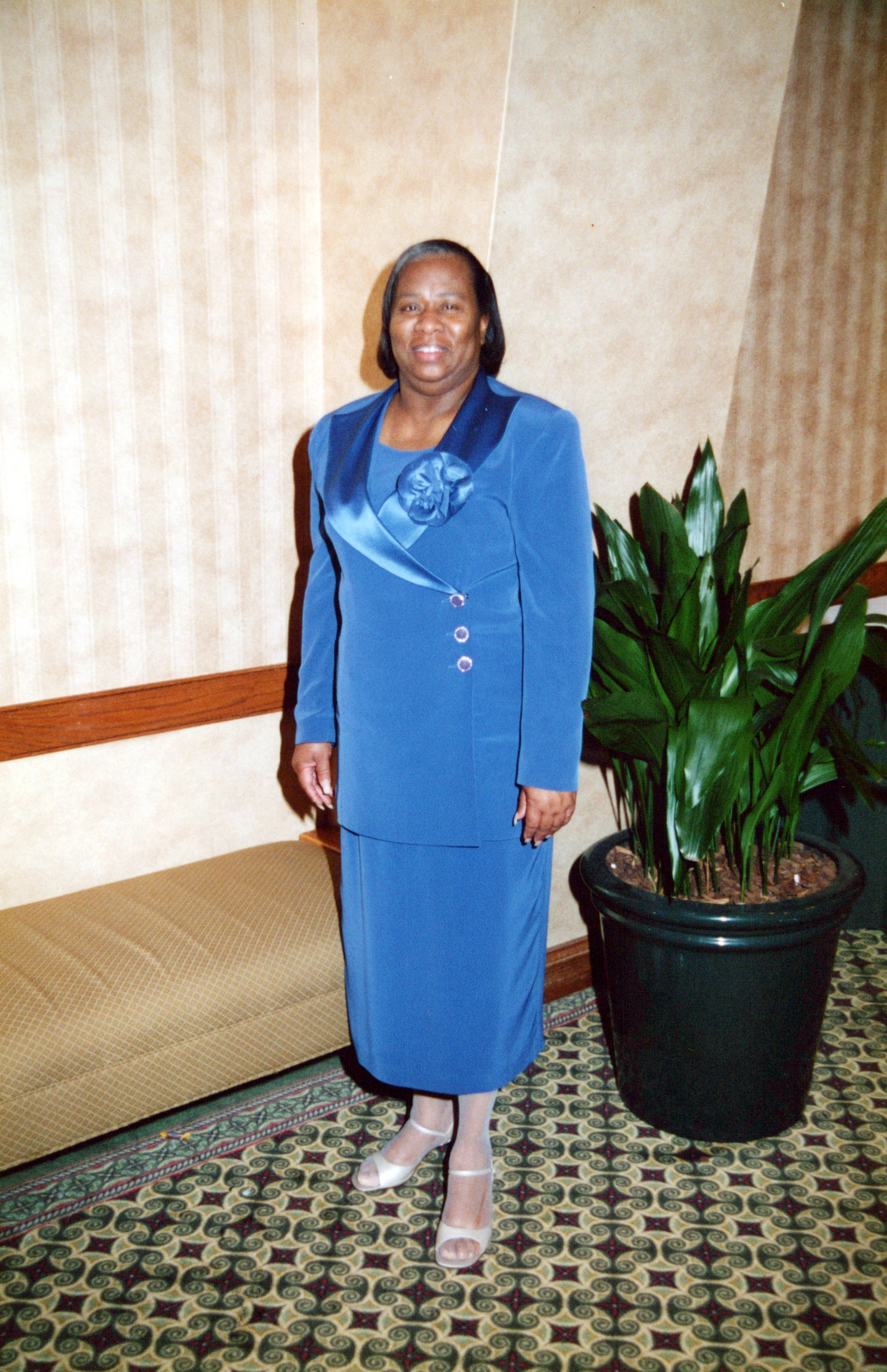 A full-length portrait of an older Black woman smiling. She wears a formal royal blue skirt suit and stands indoors next to a large potted plant.