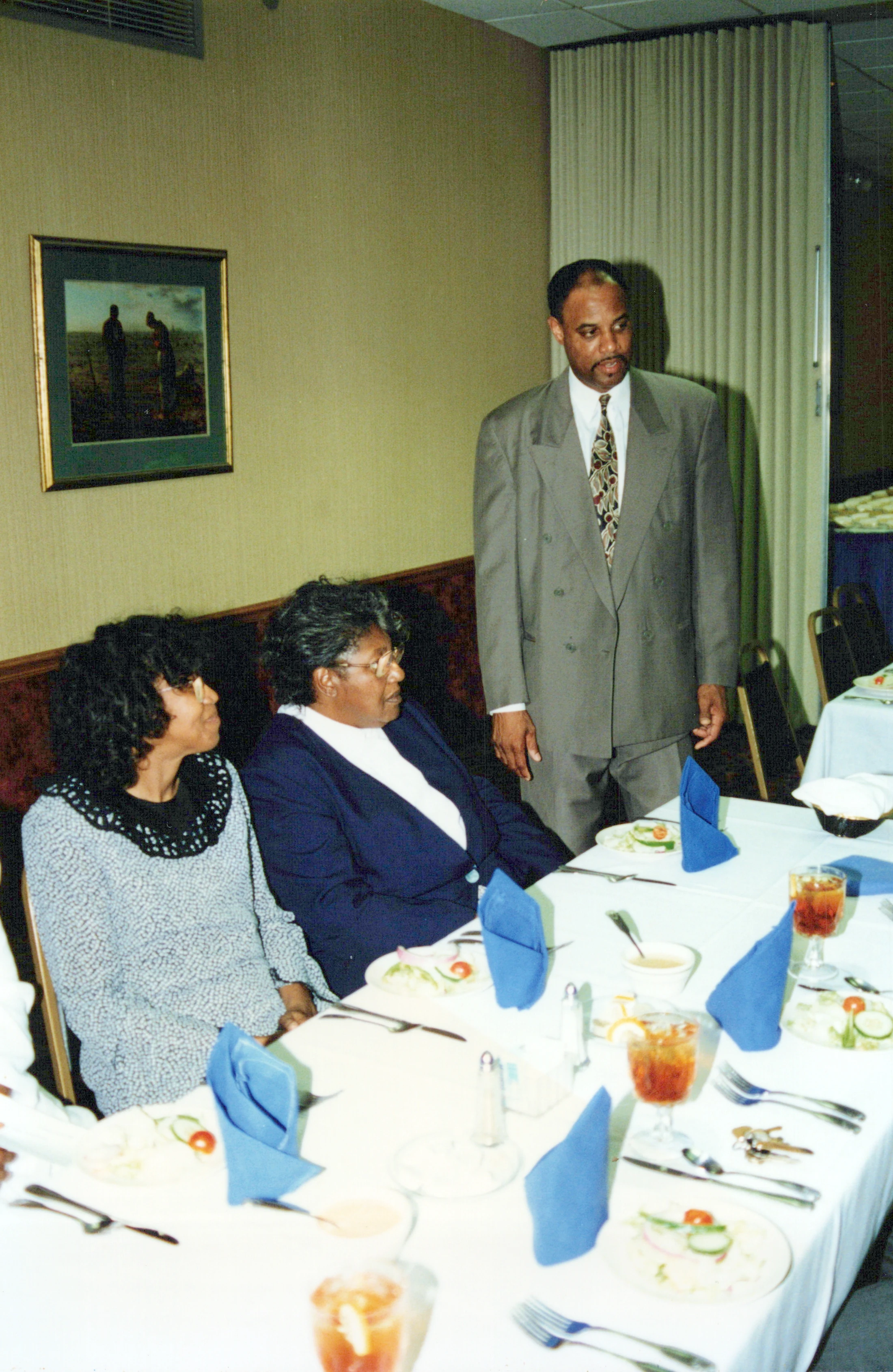 A vintage photo of two Black women sitting at a dinner table while a Black man in a suit stands behind them. The table is set with salads and blue napkins.