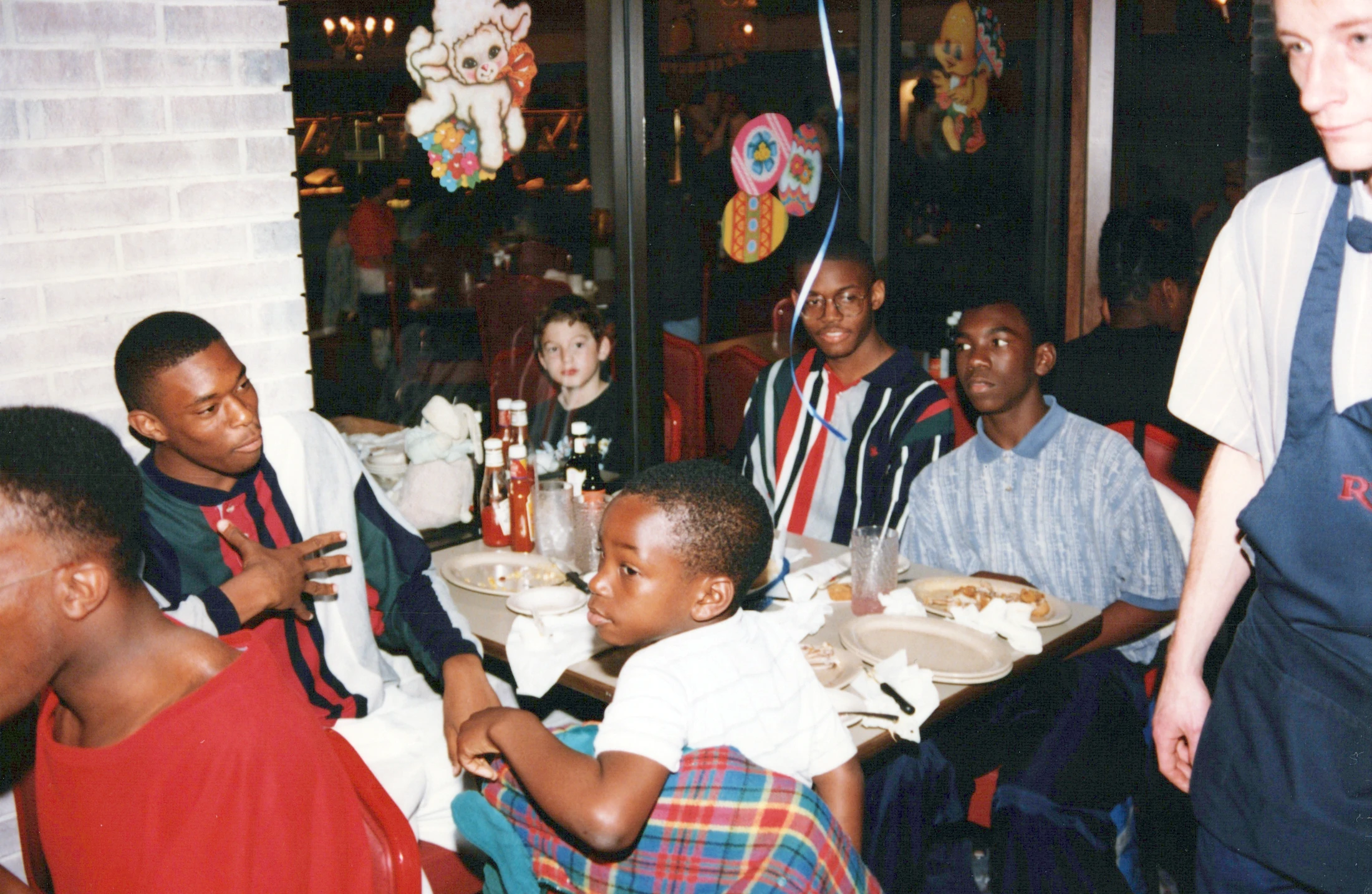 A group of young Black men and boys sit at a restaurant table with a young white boy. A waiter stands nearby. Easter decorations are on the window behind them.