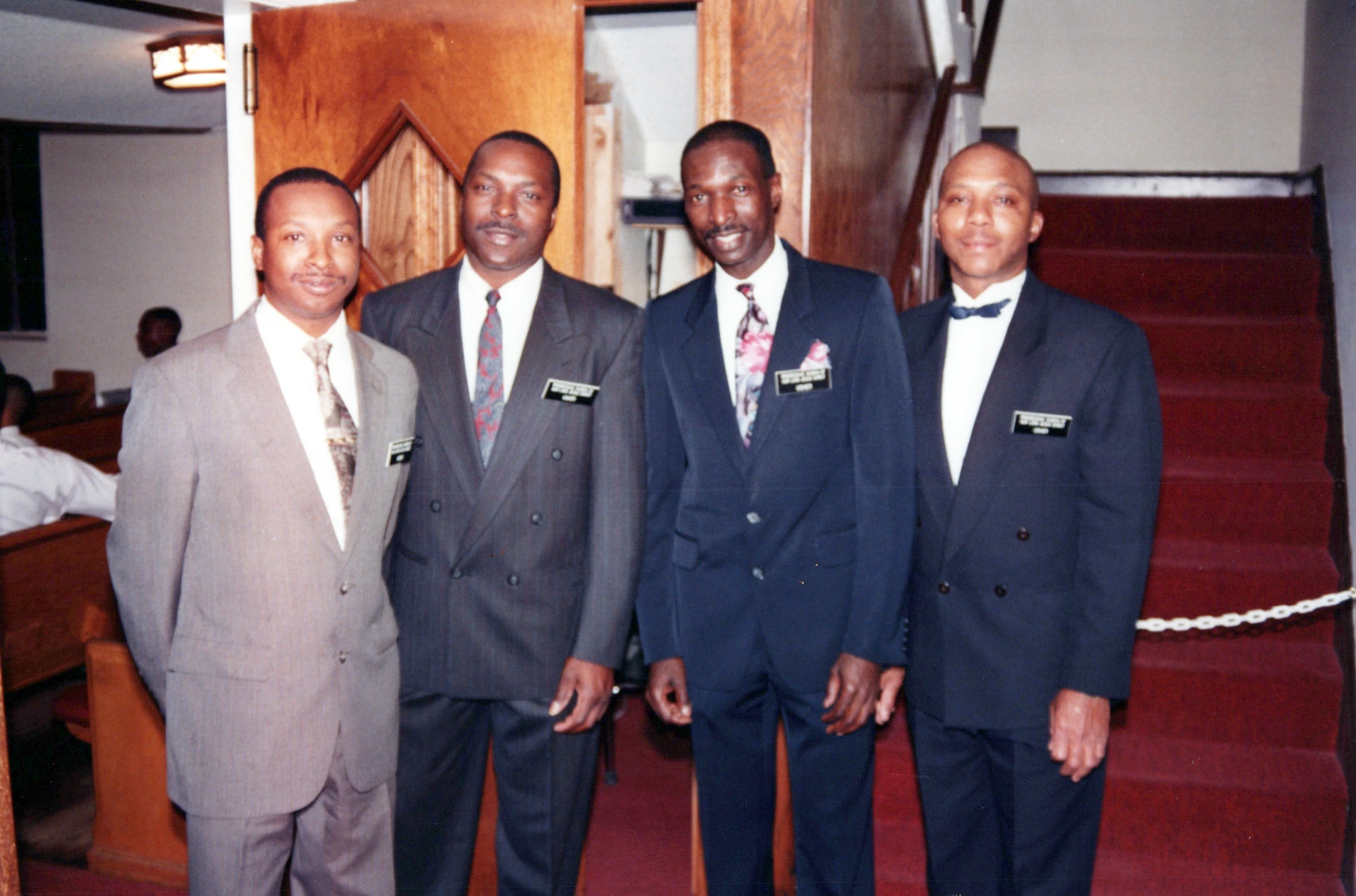 A vintage photograph of four smiling African American men in suits and ties standing together inside a church. They are wearing name tags on their lapels.