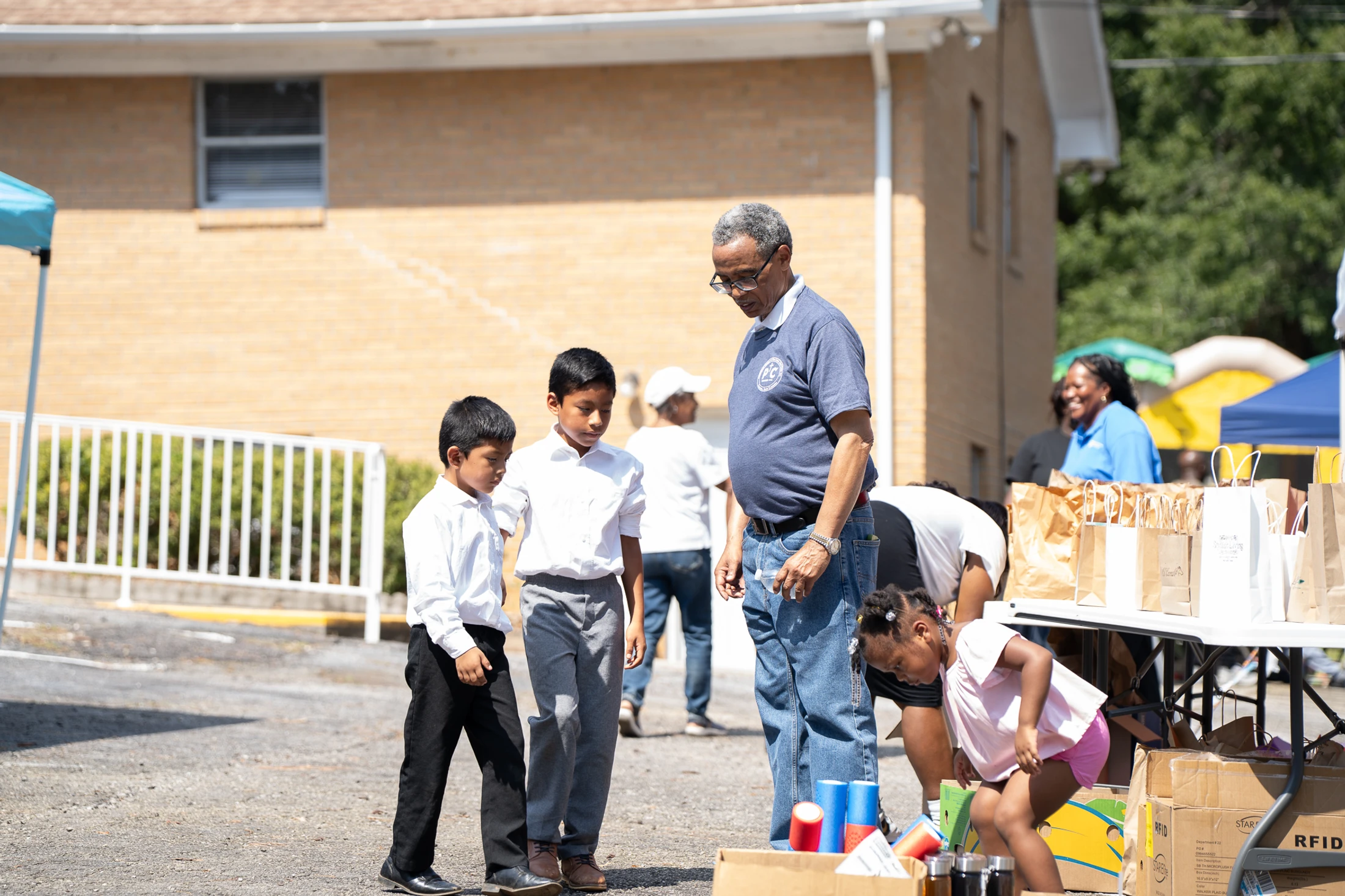 An older man talks with two young boys while a little girl looks in a box of supplies at a sunny outdoor community event with giveaway bags on a table.