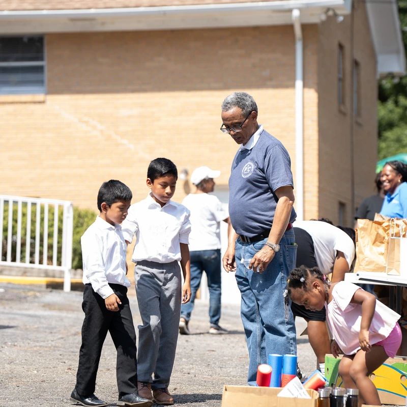 An older man talks with two young boys while a little girl looks in a box of supplies at a sunny outdoor community event with giveaway bags on a table.