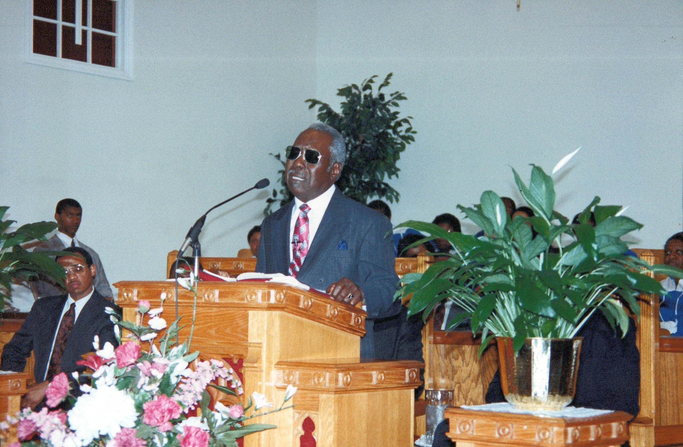 An older Black man in a suit and sunglasses stands at a wooden pulpit in a church, speaking into a microphone during a service.