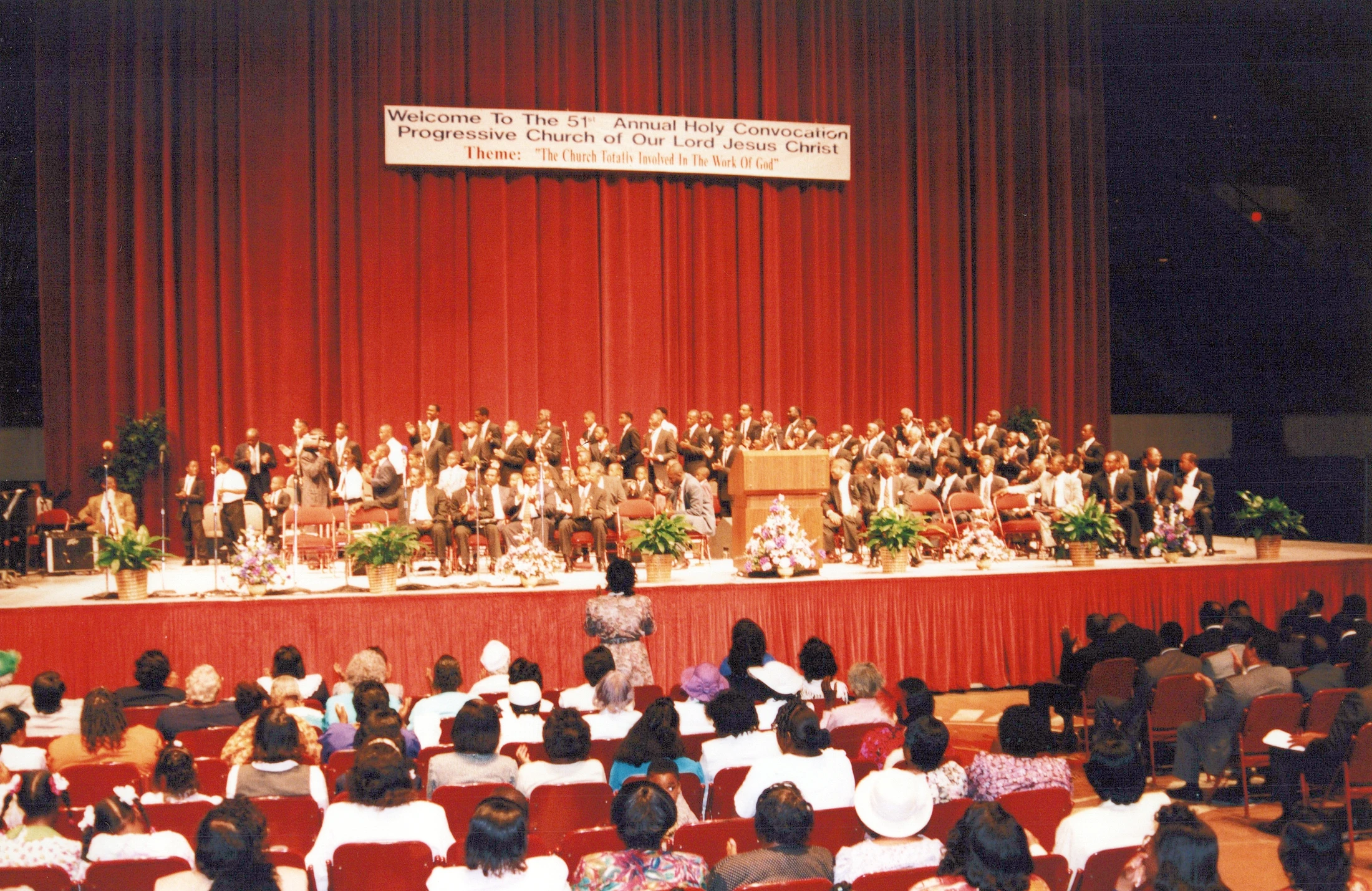 A view from the audience of a church convocation. Men in suits sit on a stage in front of a large red curtain with a banner welcoming attendees.