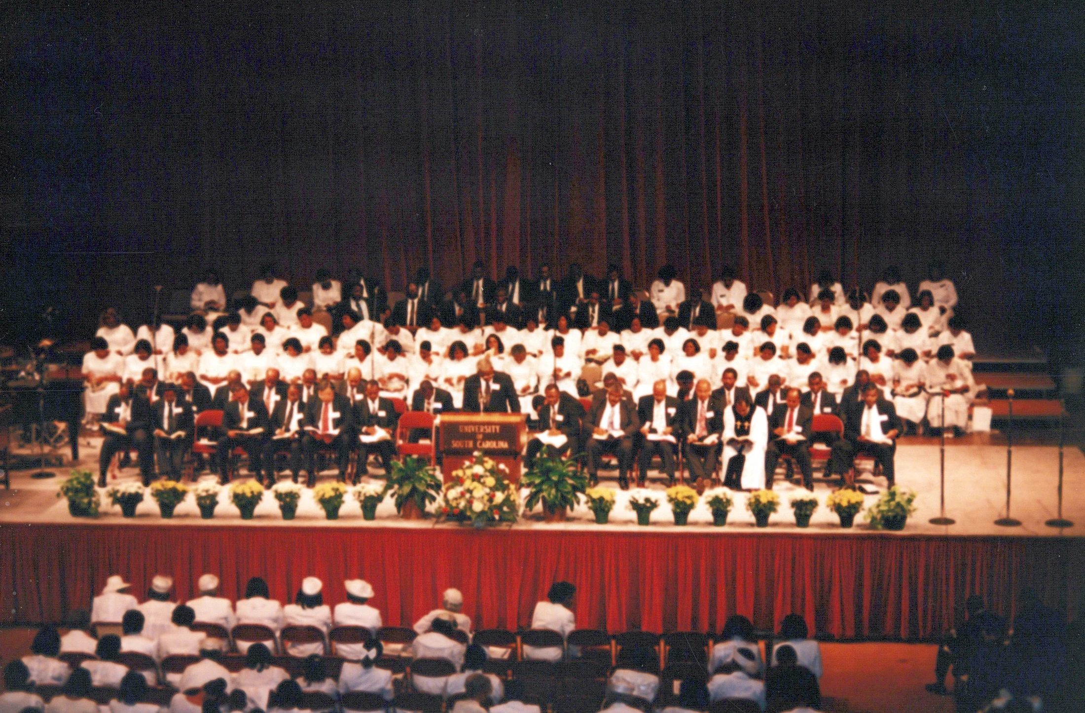 A wide shot of a large group on an auditorium stage for a University of South Carolina event. Men in suits sit in front of a choir of women in white.