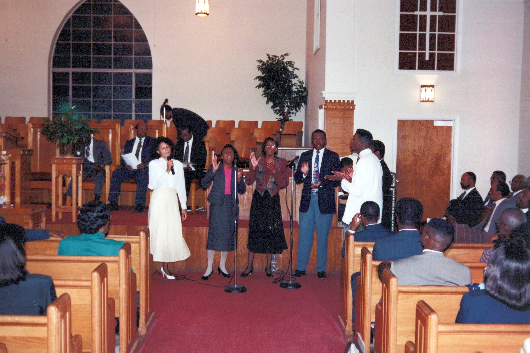 A group of Black men and women sing joyfully at the front of a church. A congregation watches from wooden pews during a worship service.
