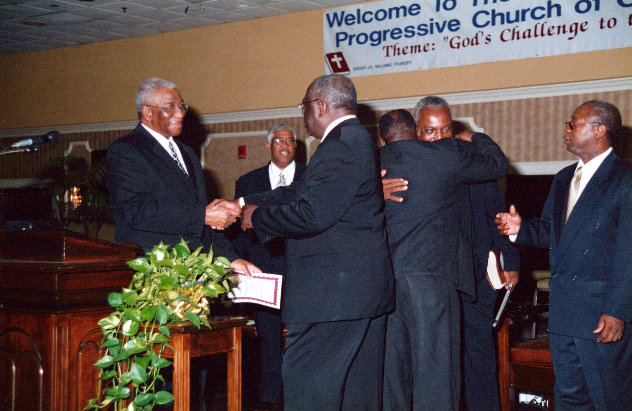 A group of older Black men in suits at a church function. A man at a lectern shakes another's hand, while other men embrace in the background.
