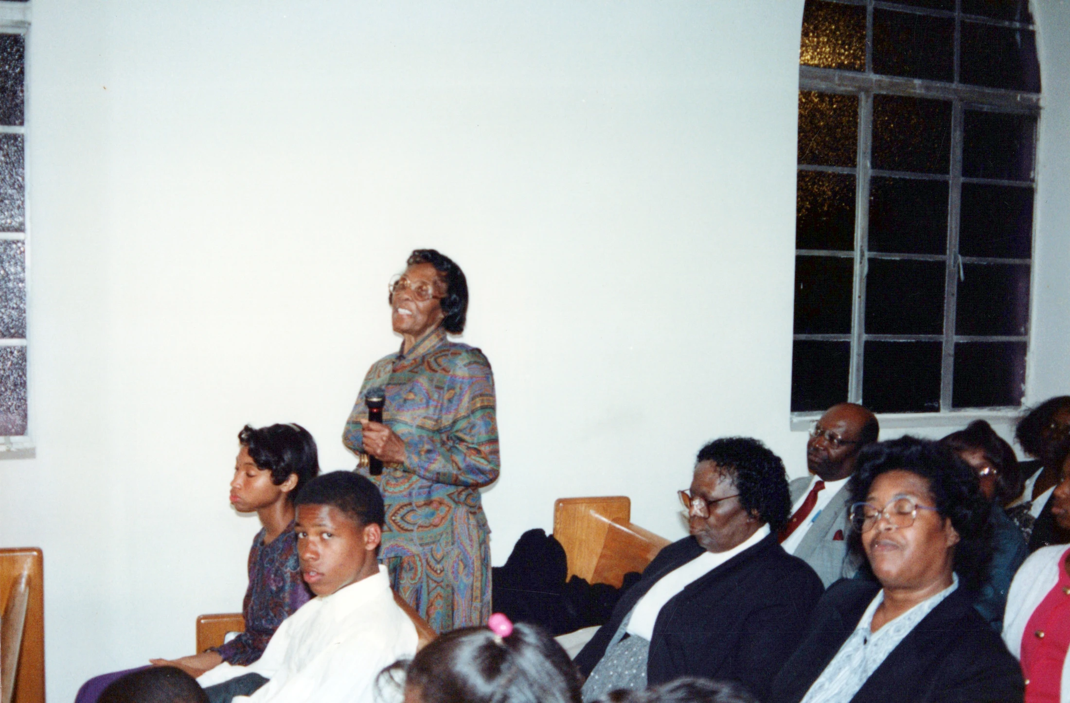 An elderly Black woman in a colorful dress stands and speaks into a microphone while a congregation of Black men, women, and children watch from wooden pews.