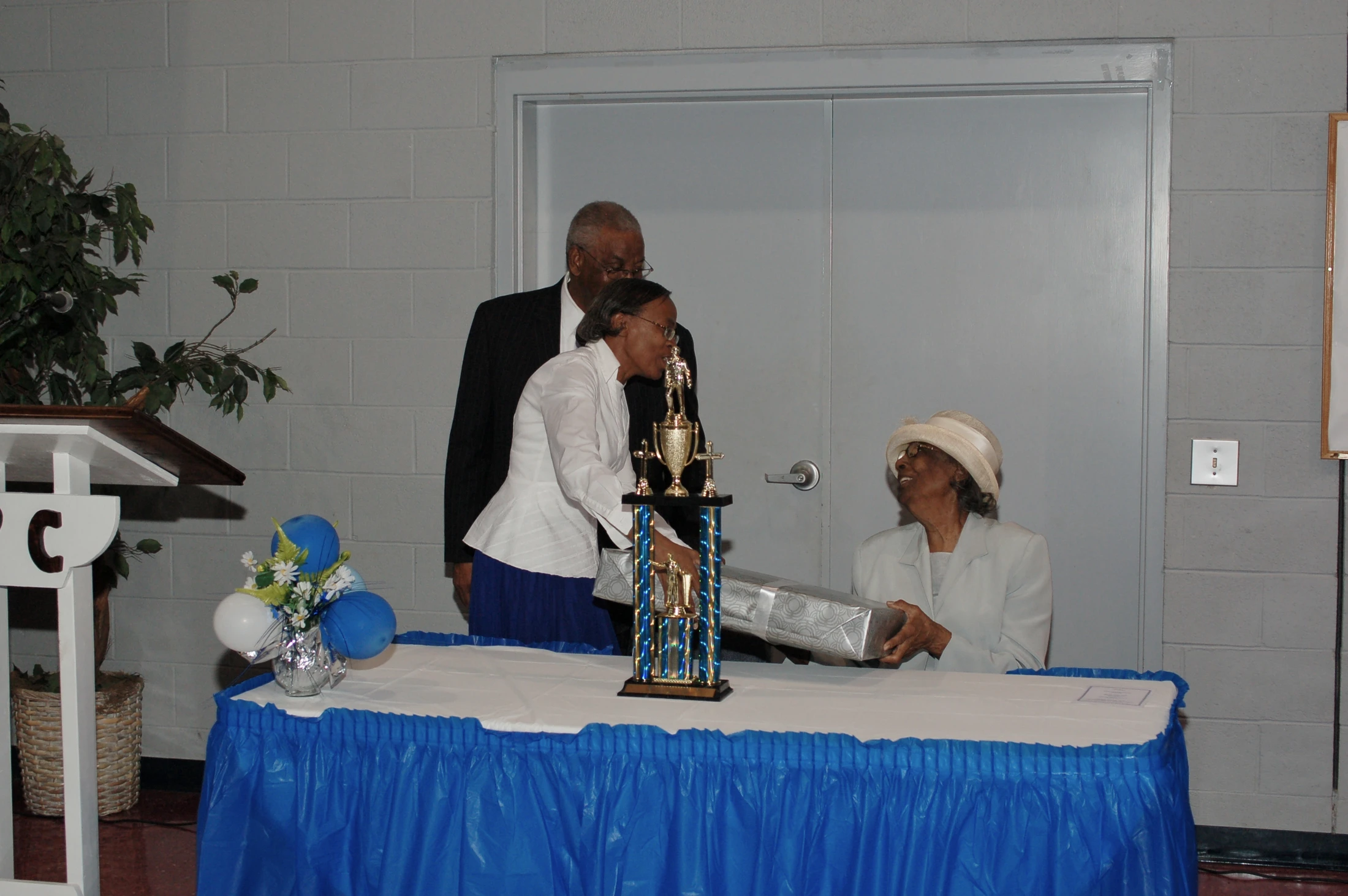 A woman in a white shirt presents a wrapped gift to a smiling older woman in a hat, sitting at a table with a trophy. A man in a suit stands behind them.