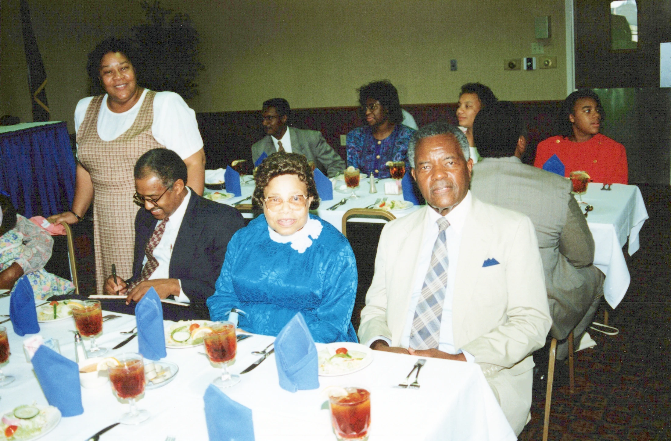 A vintage photograph showing a group of formally dressed African American guests enjoying a meal at a banquet, seated around tables with white tablecloths.