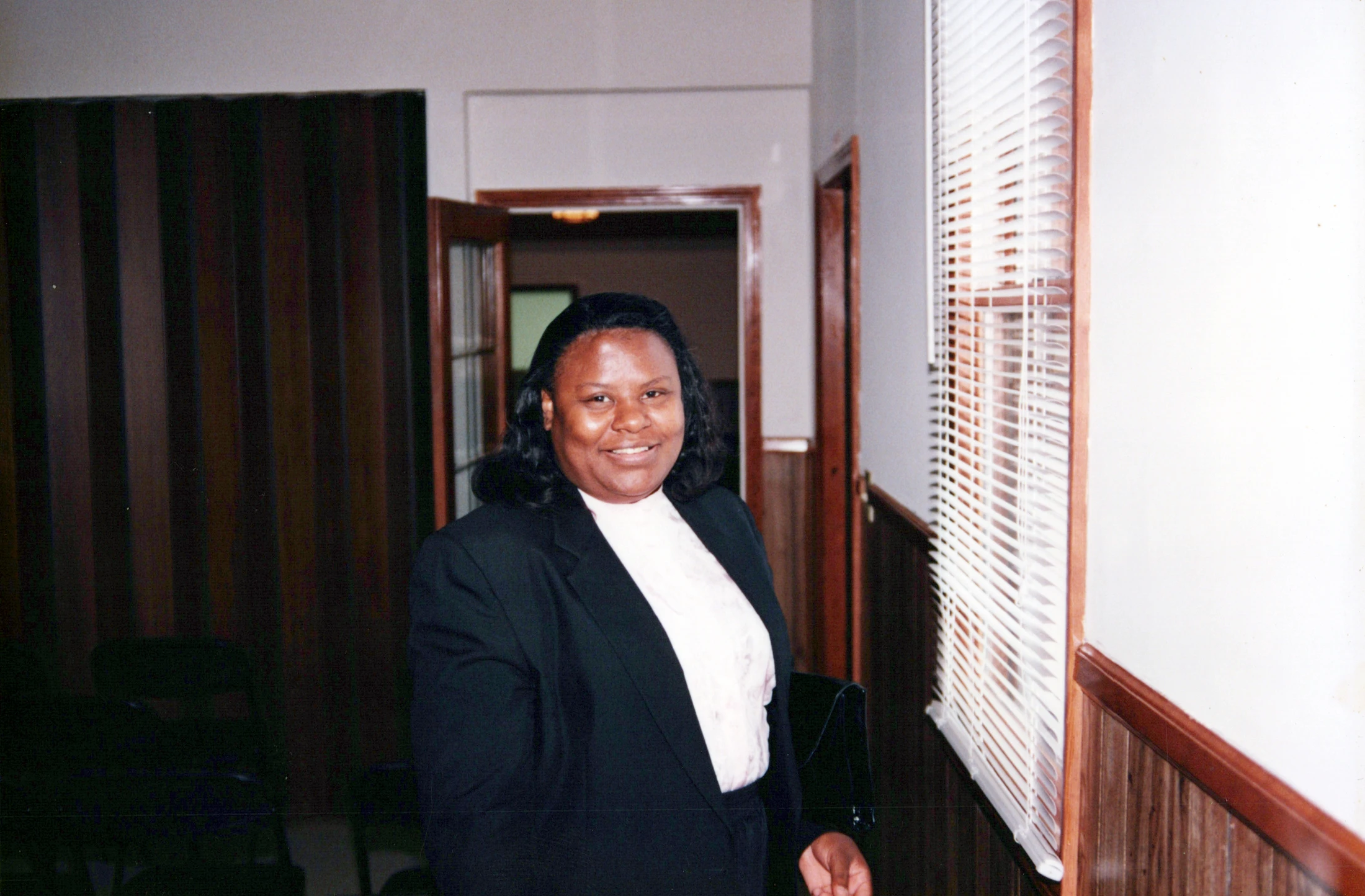A Black woman with shoulder-length dark hair smiles at the camera. She wears a black blazer and a white blouse, standing indoors near a window with blinds.