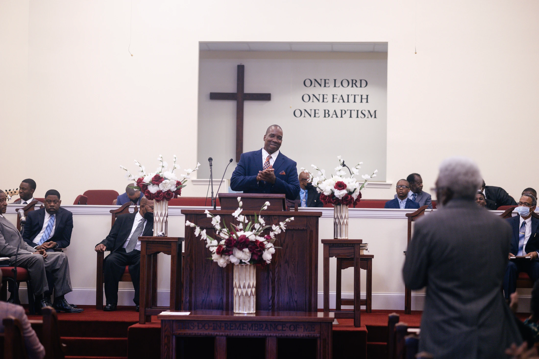 A smiling Black pastor in a suit stands at a wooden pulpit in a church. Behind him on the wall is a cross and the words 'One Lord One Faith One Baptism'.