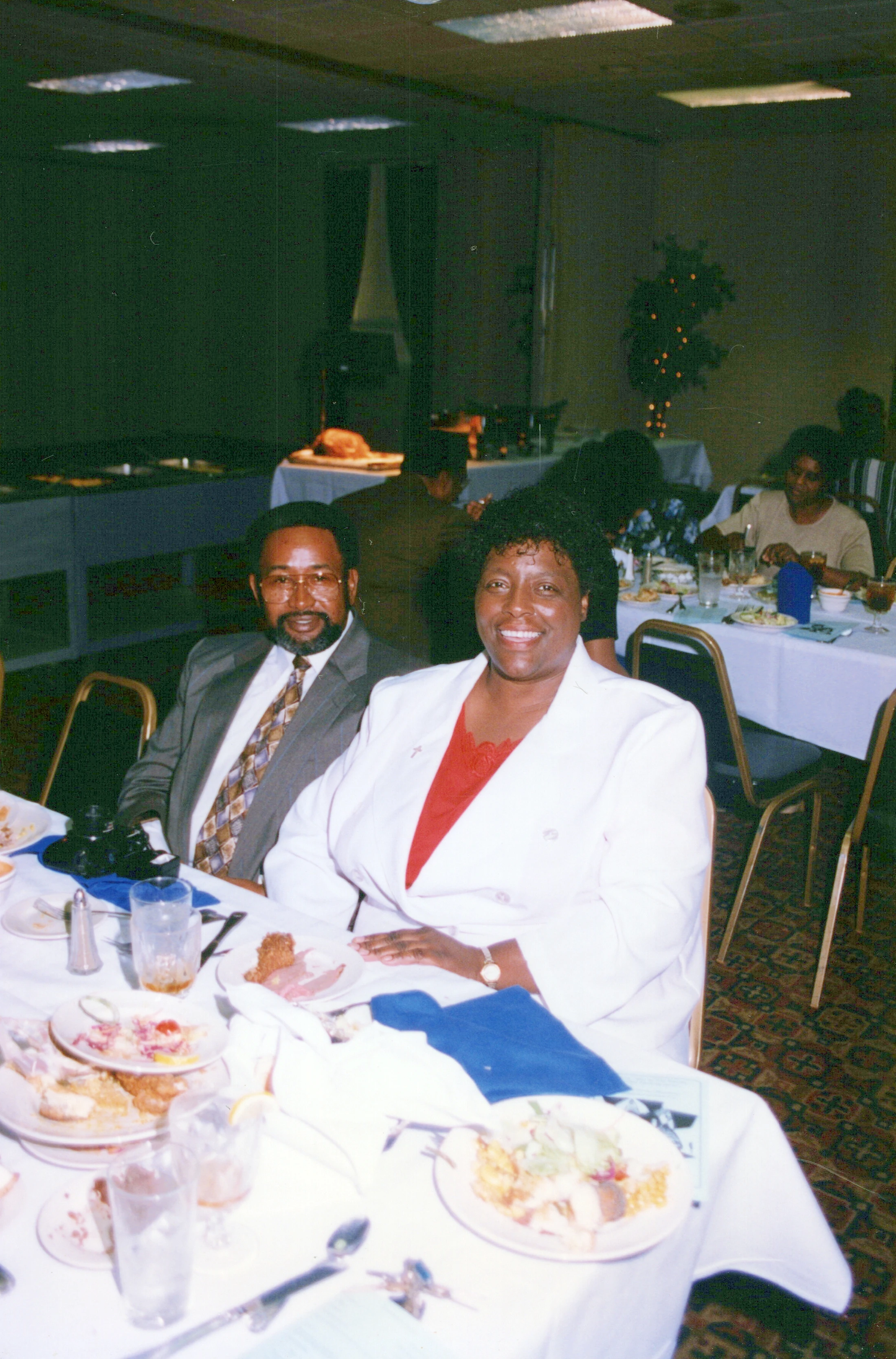 A vintage film photo of a smiling Black woman in a white blazer and a man in a suit sitting together at a messy dinner table during an indoor event.