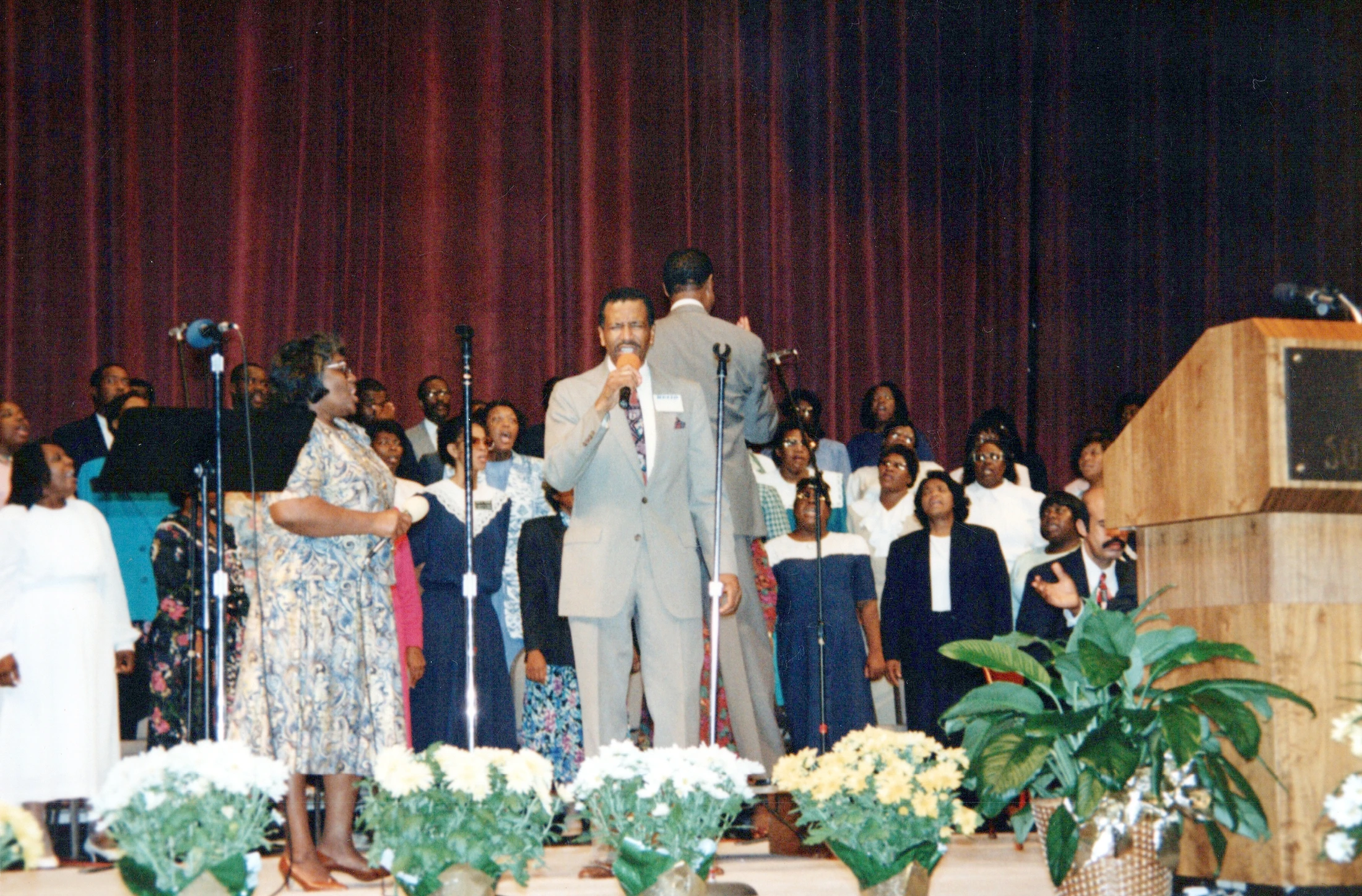 A vintage photo of a Black church choir on stage. A man in a light-colored suit holds a microphone and sings, with other members singing behind him.