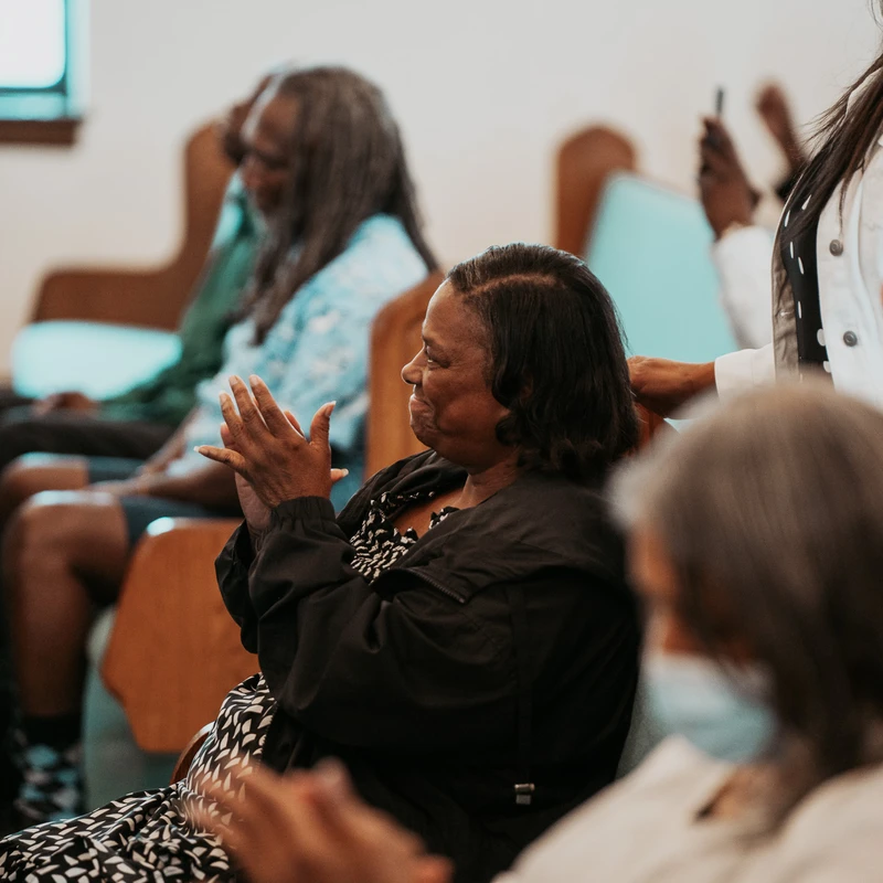 A Black woman with a gentle smile sits in a church pew, clapping her hands. Other members of the congregation are seated around her in the pews.