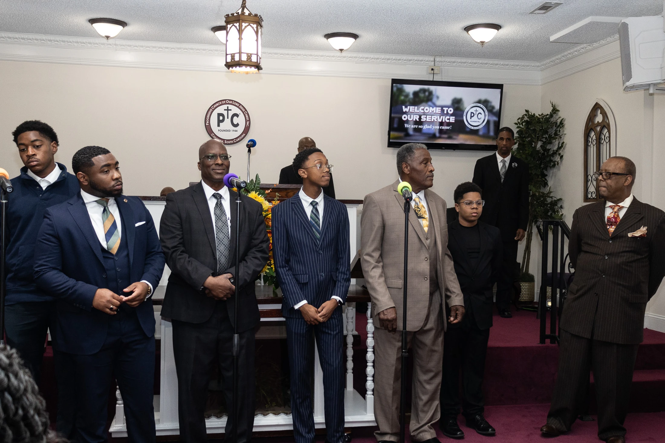 A group of Black men and boys of various ages, dressed in suits, stand at the front of a church sanctuary during a service, with microphones before them.