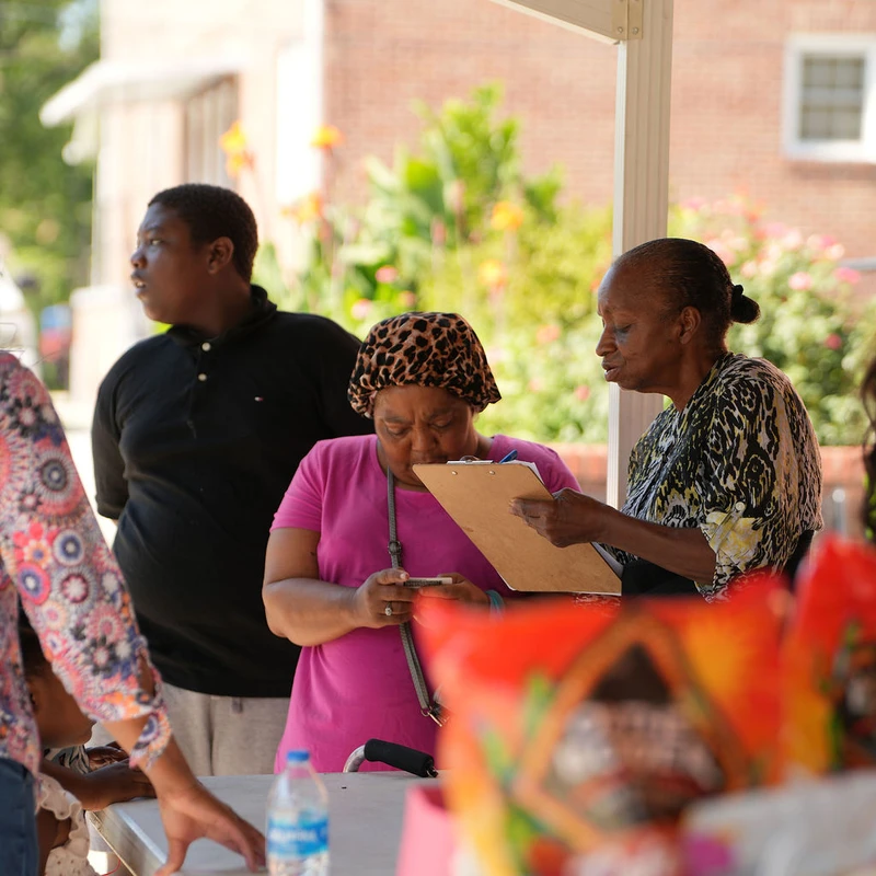 Three people stand at a table outdoors. A woman in a pink shirt and another woman look at a clipboard, while a young man looks on. Bags of snacks are blurred.