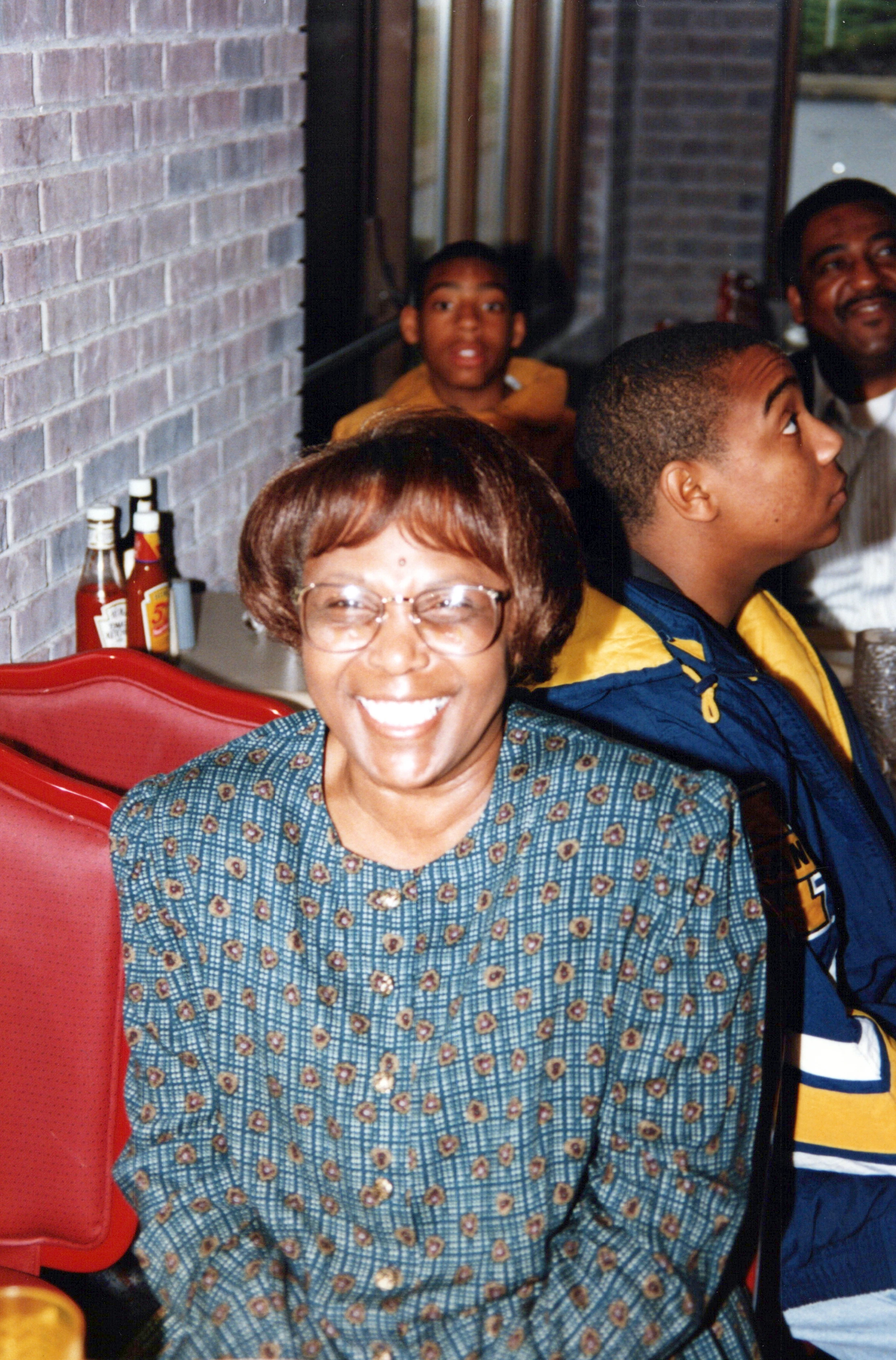 A smiling Black woman wearing glasses and a patterned blouse sits in a red booth. A young man sits beside her, and two other people are in the background.