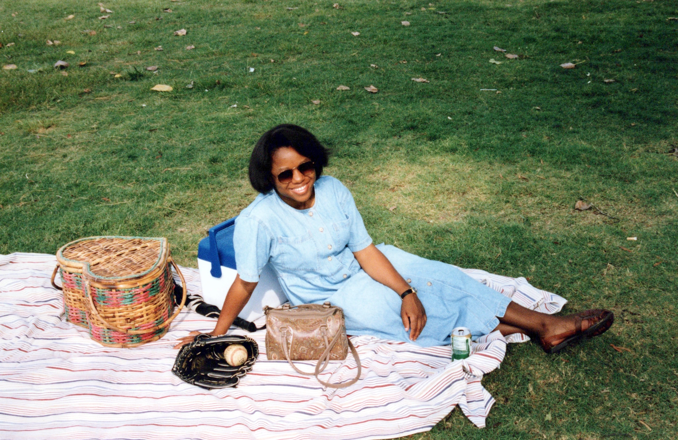 A smiling woman in a blue dress and sunglasses sits on a striped blanket in a grassy park, next to a picnic basket and a baseball glove with a ball.