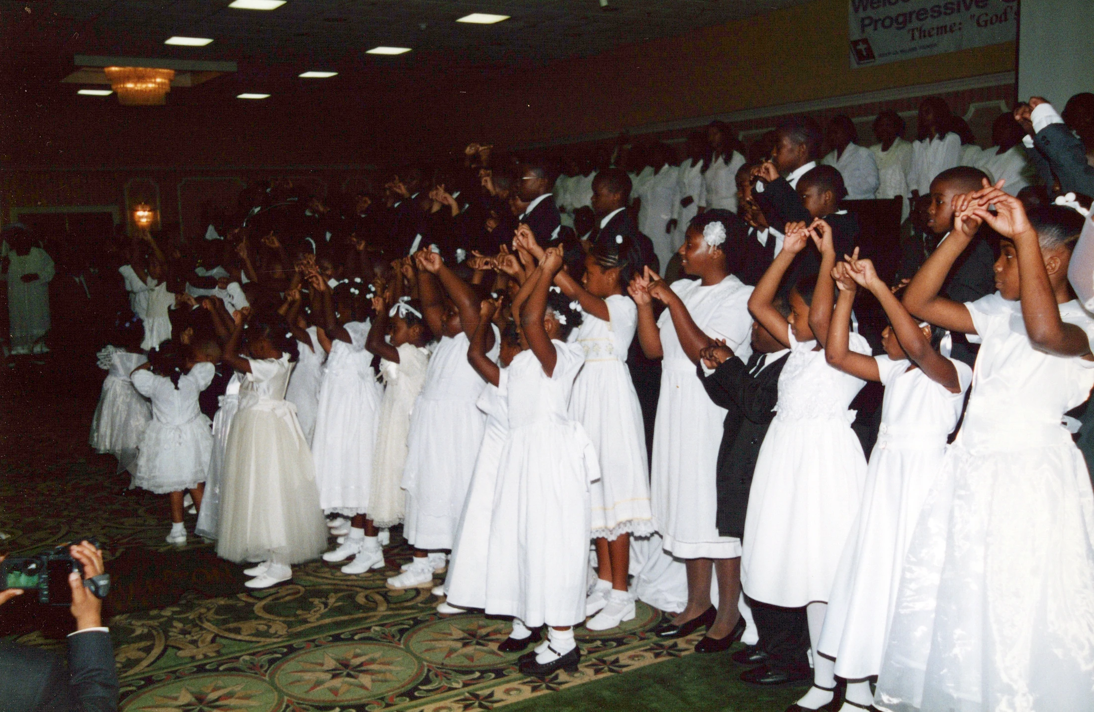 A large group of young Black children, girls in white dresses and boys in suits, stand together in a hall with their arms raised during a church service.