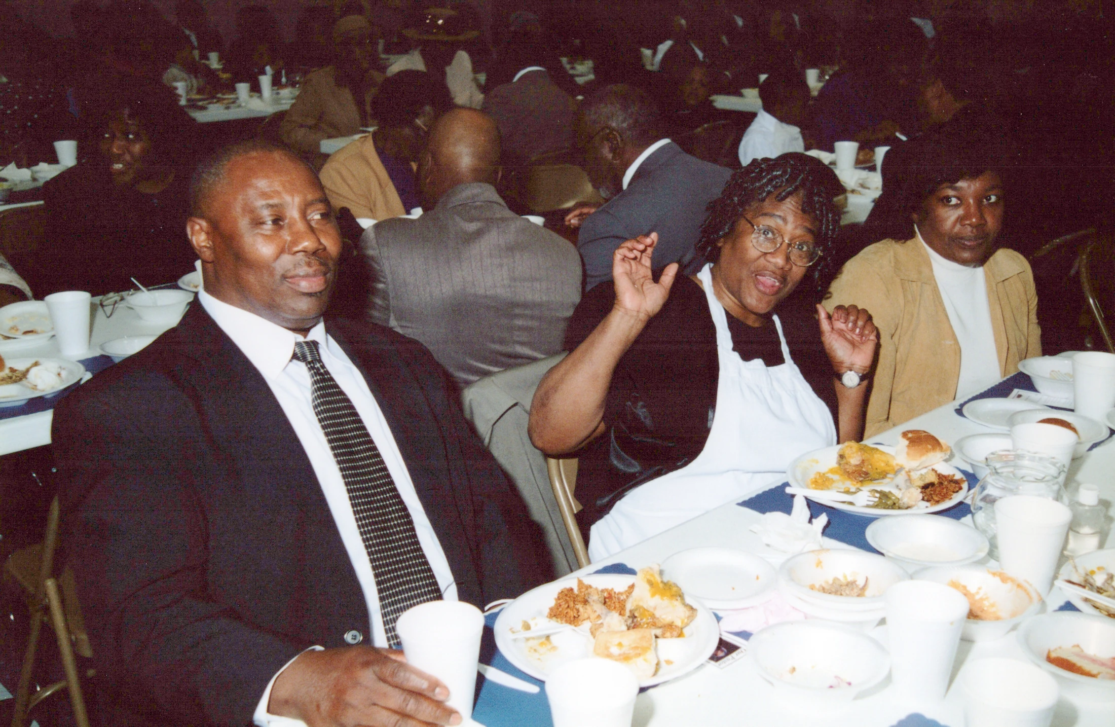 A group of Black adults enjoying a meal at a banquet. A man in a suit sits next to a smiling woman in an apron who is waving at the camera.