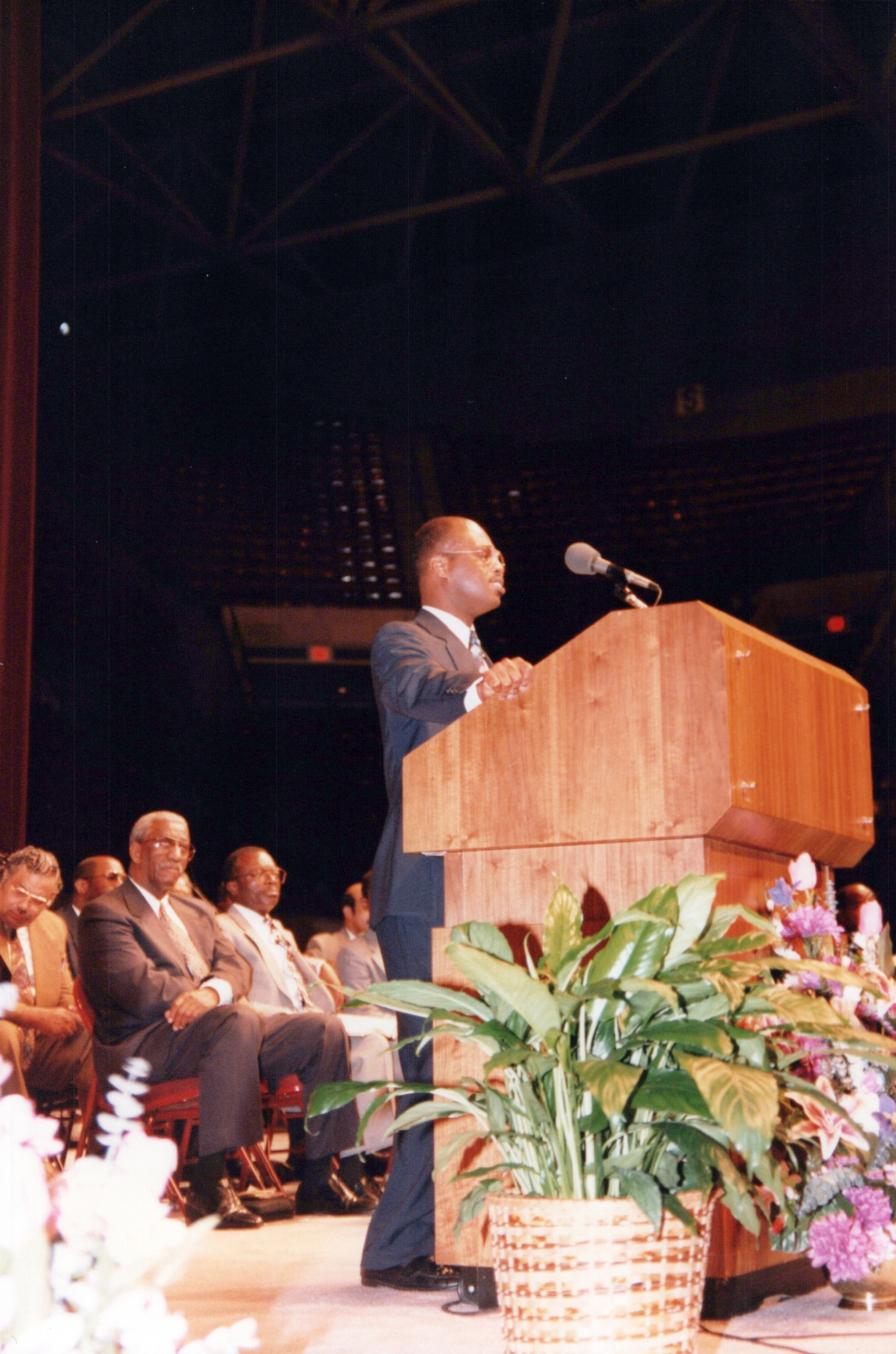 A man in a dark suit and glasses stands at a wooden podium speaking into a microphone. In the background, other men are seated on the stage in a large hall.