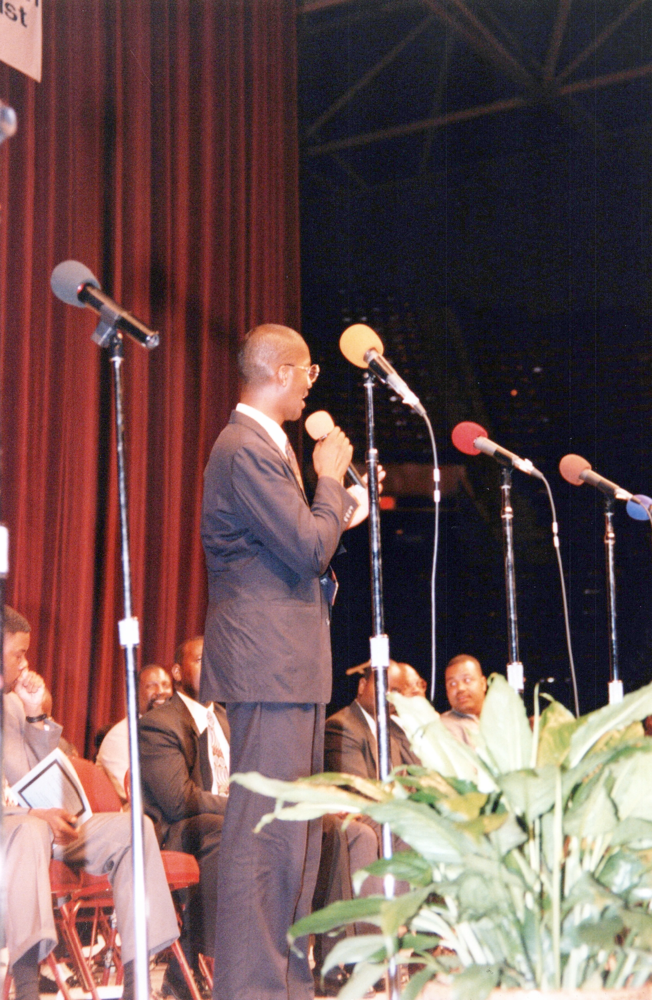 A Black man in a suit and glasses stands on a stage with a red curtain, speaking into a microphone. Other men are seated listening in the background.