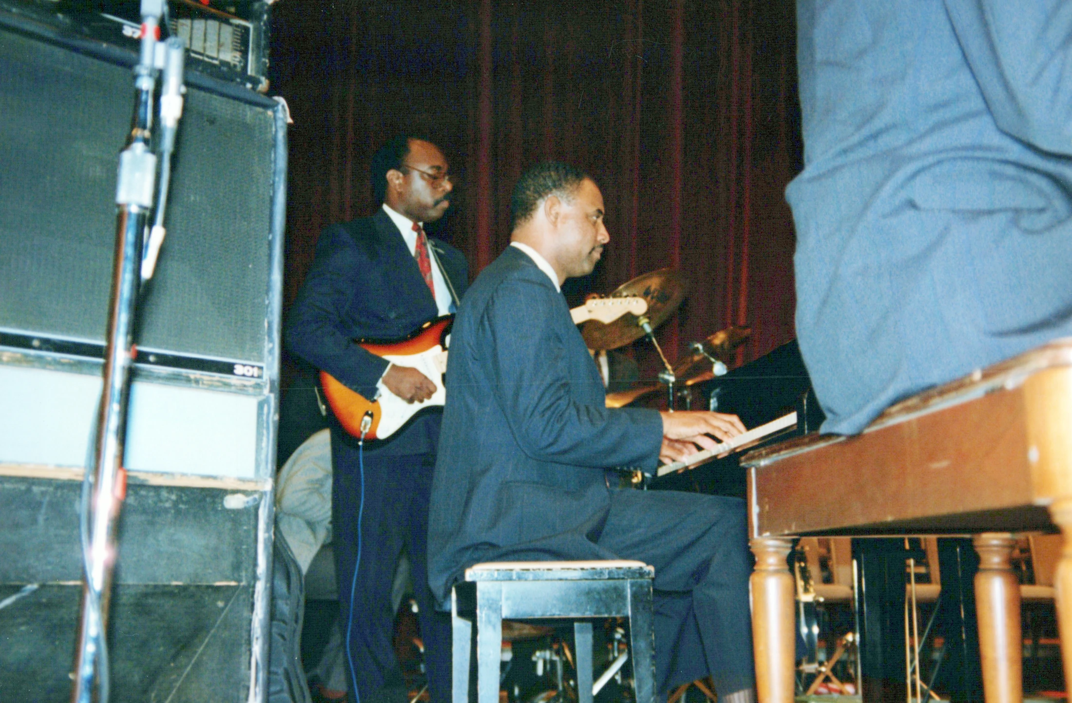 A man in a dark suit plays the piano on stage, while another man in a suit stands behind him playing an electric guitar in front of a red curtain.