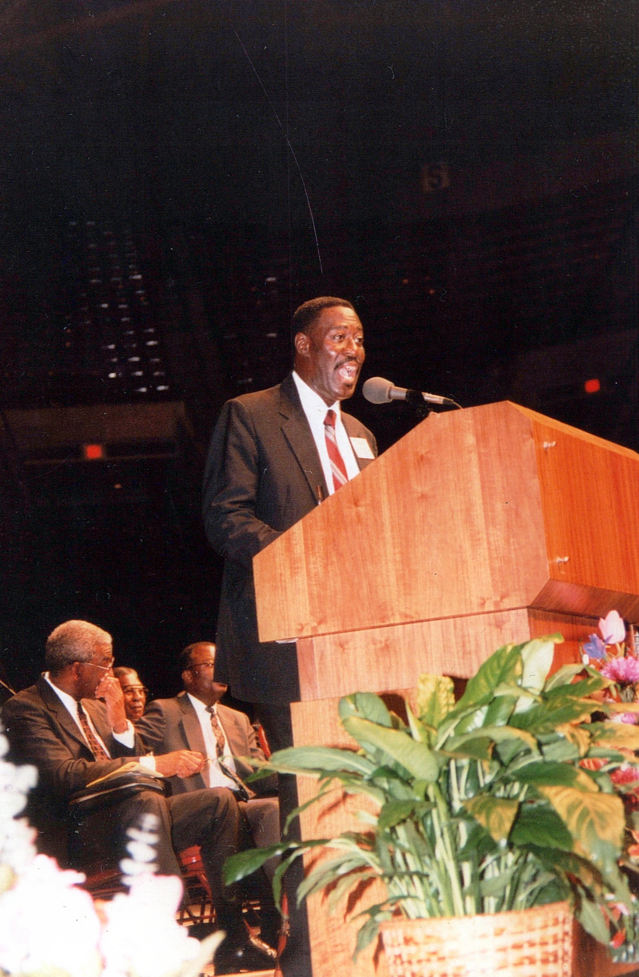 A smiling Black man in a dark suit and red tie speaks into a microphone at a wooden lectern on a stage. Other men are seated behind him.