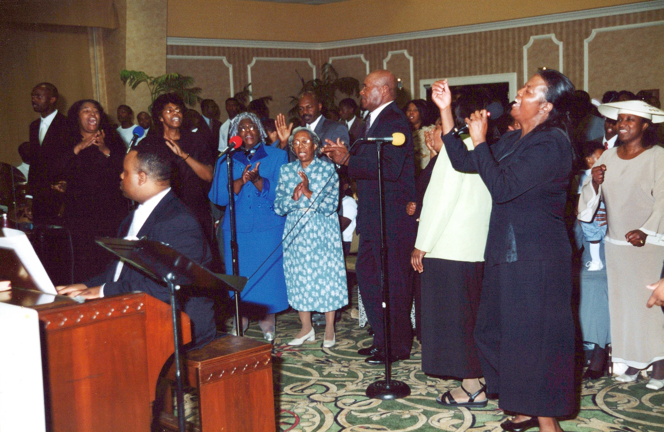 A large, formally dressed African American choir sings with great emotion. A man plays an organ while others clap, raise their hands, and sing into microphones.