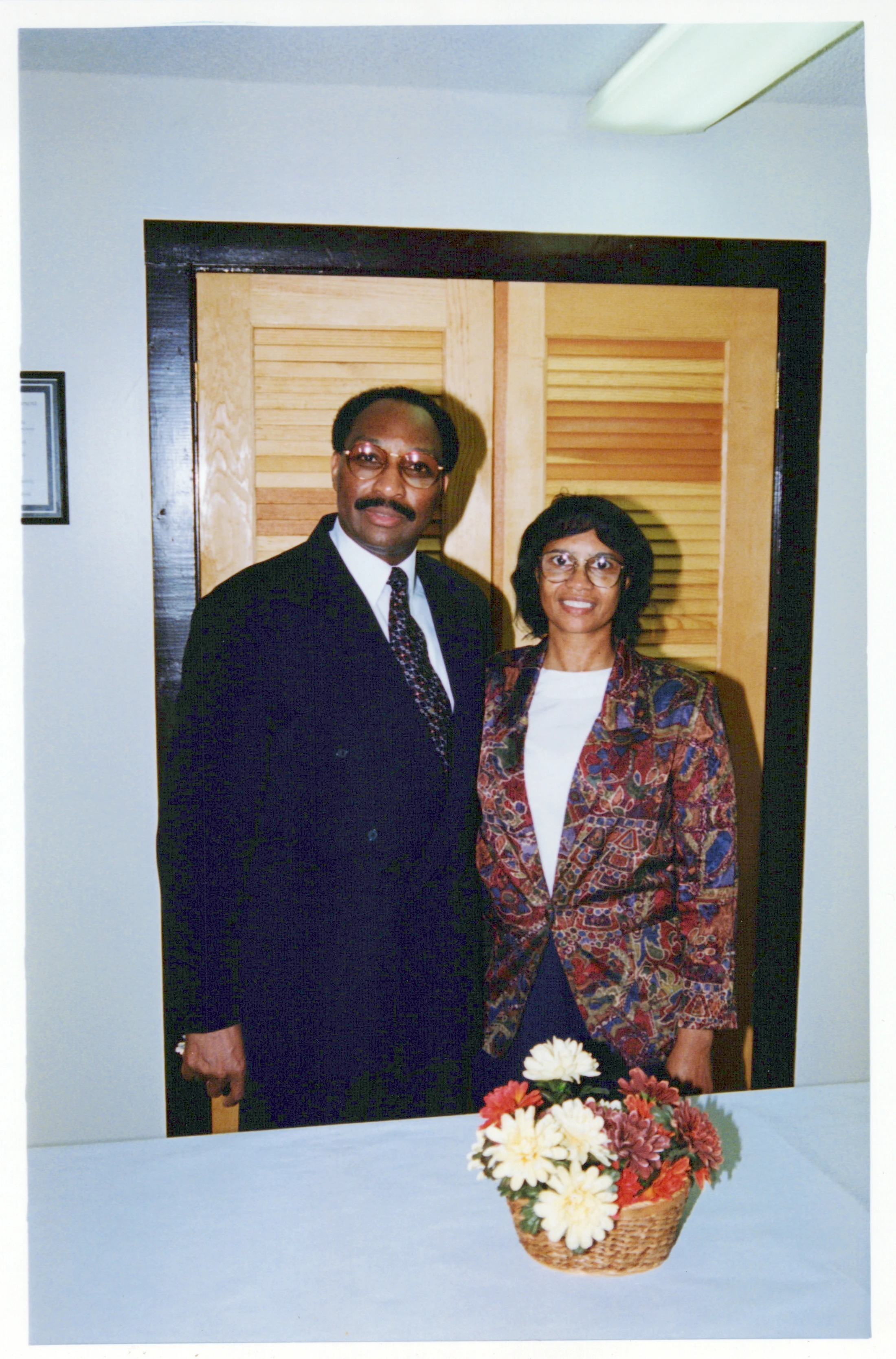 An African American man in a suit and a woman in a patterned blazer stand together behind a table with a basket of flowers. They are smiling at the camera.
