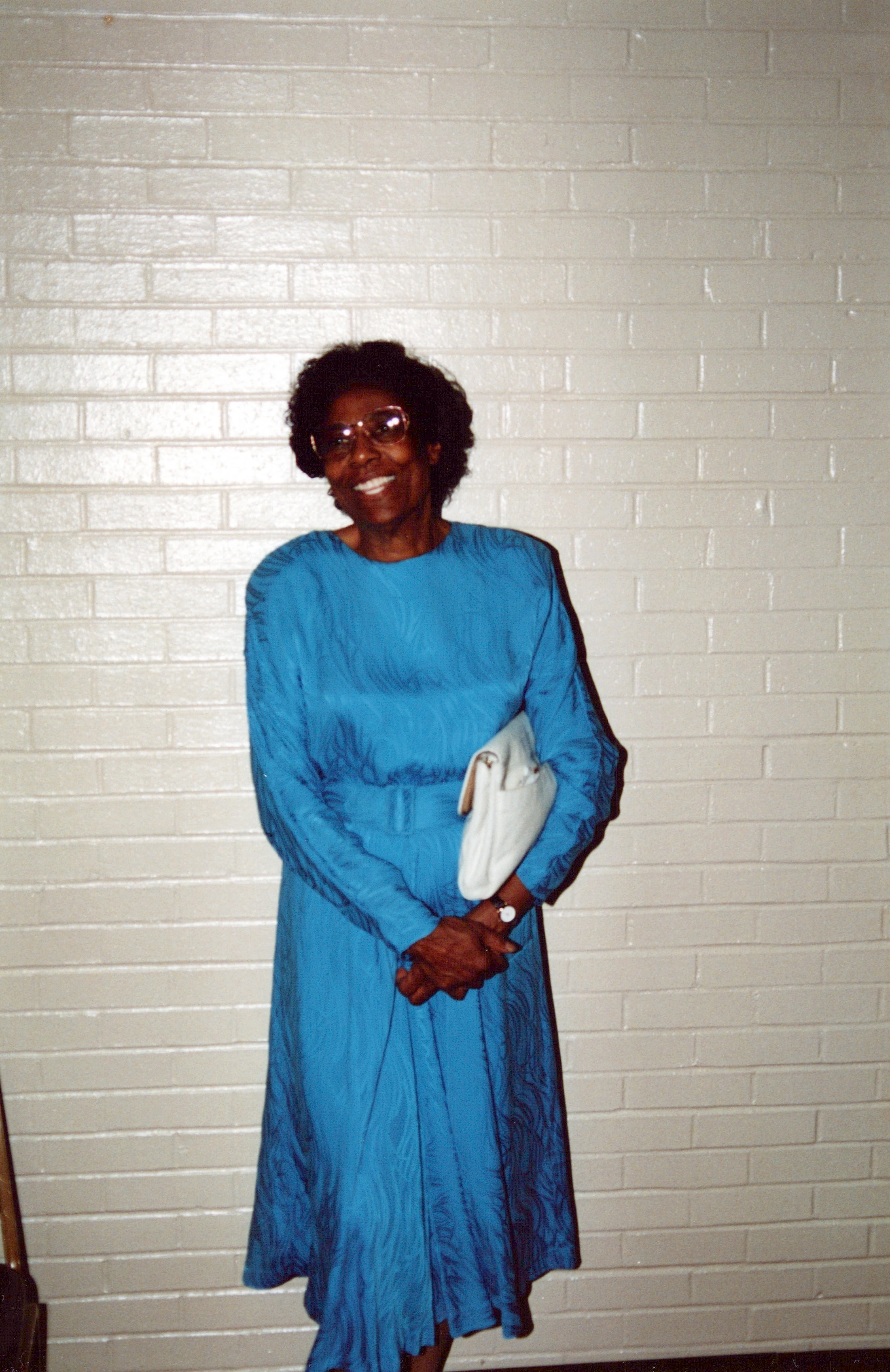 An older Black woman with glasses and short hair smiles in a vibrant blue dress. She stands against a white brick wall, holding a white clutch purse.