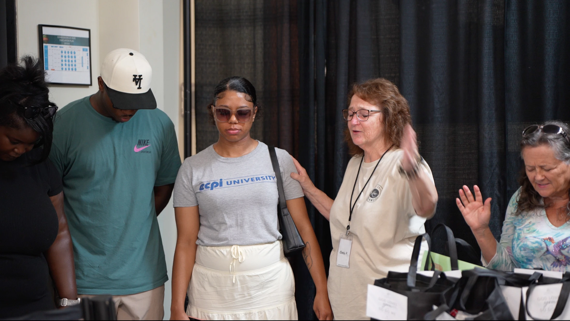 An older woman with glasses leads a diverse group of people in prayer, placing a hand on the shoulder of a young woman with her eyes closed.