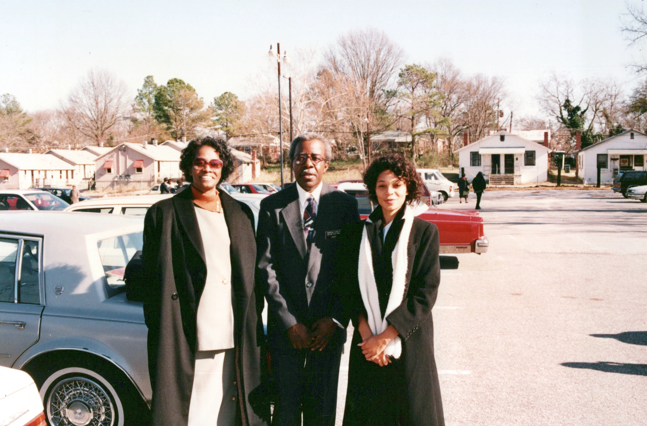 A vintage photograph of a man and two women, all Black, dressed in coats and a suit, posing together in a parking lot on a sunny day.