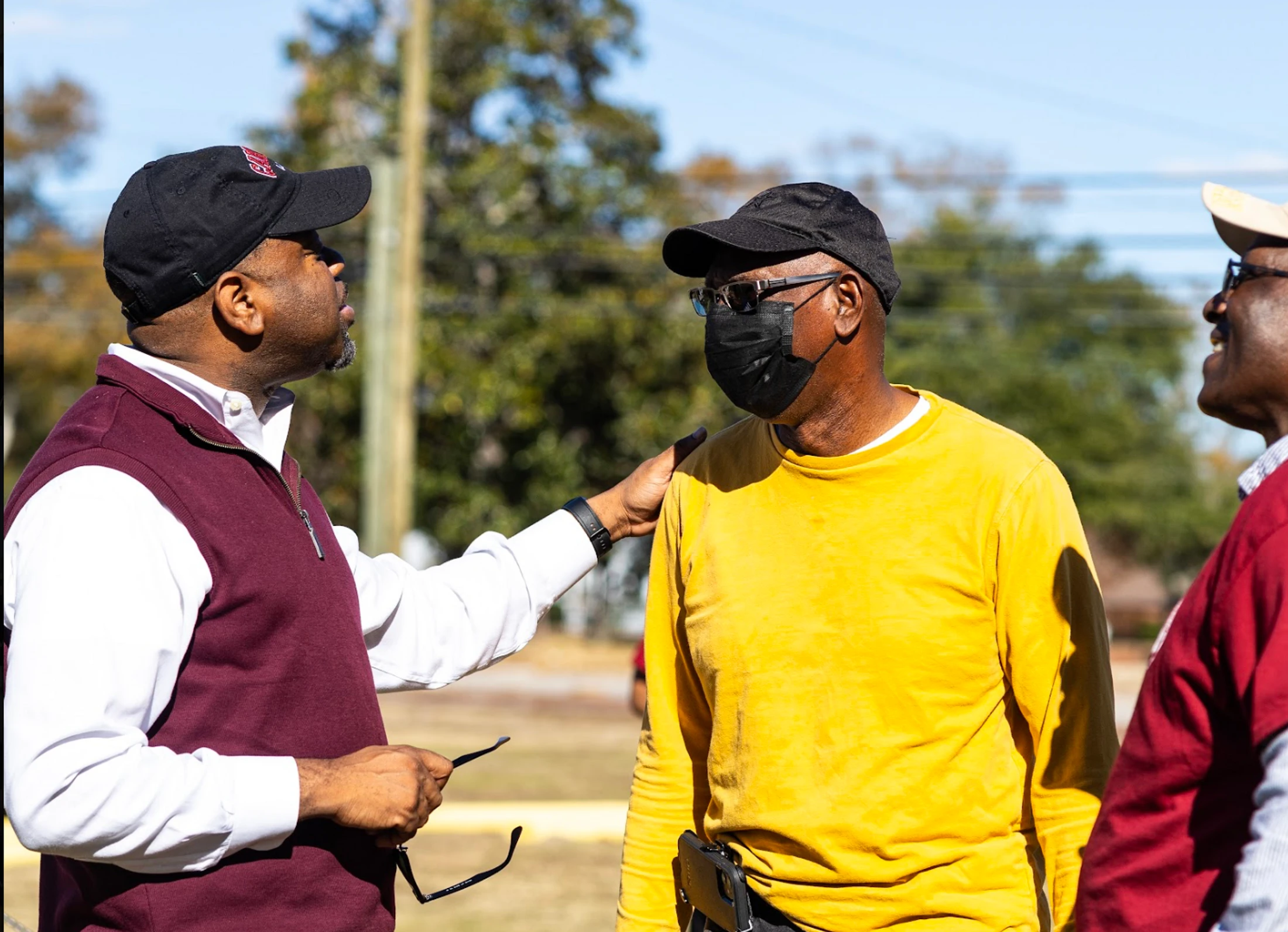 Three Black men talking outdoors. One man in a maroon vest has his hand on the shoulder of a man in a yellow shirt and face mask.
