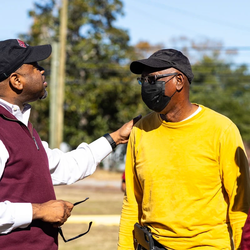 Three Black men talking outdoors. One man in a maroon vest has his hand on the shoulder of a man in a yellow shirt and face mask.