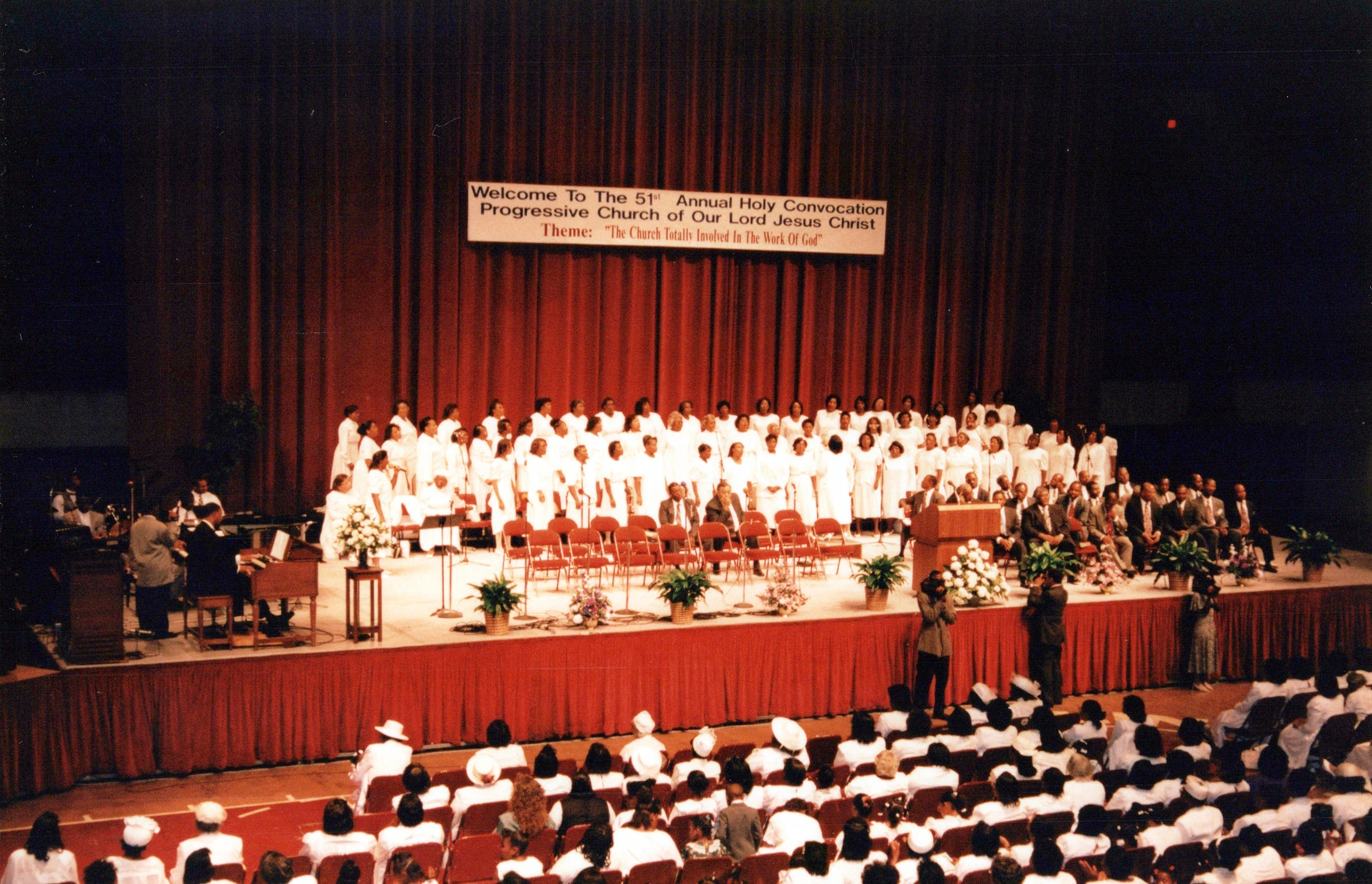 A wide shot of a church convocation with a large choir in white robes on stage, in front of a red curtain and a banner, facing a large seated audience.