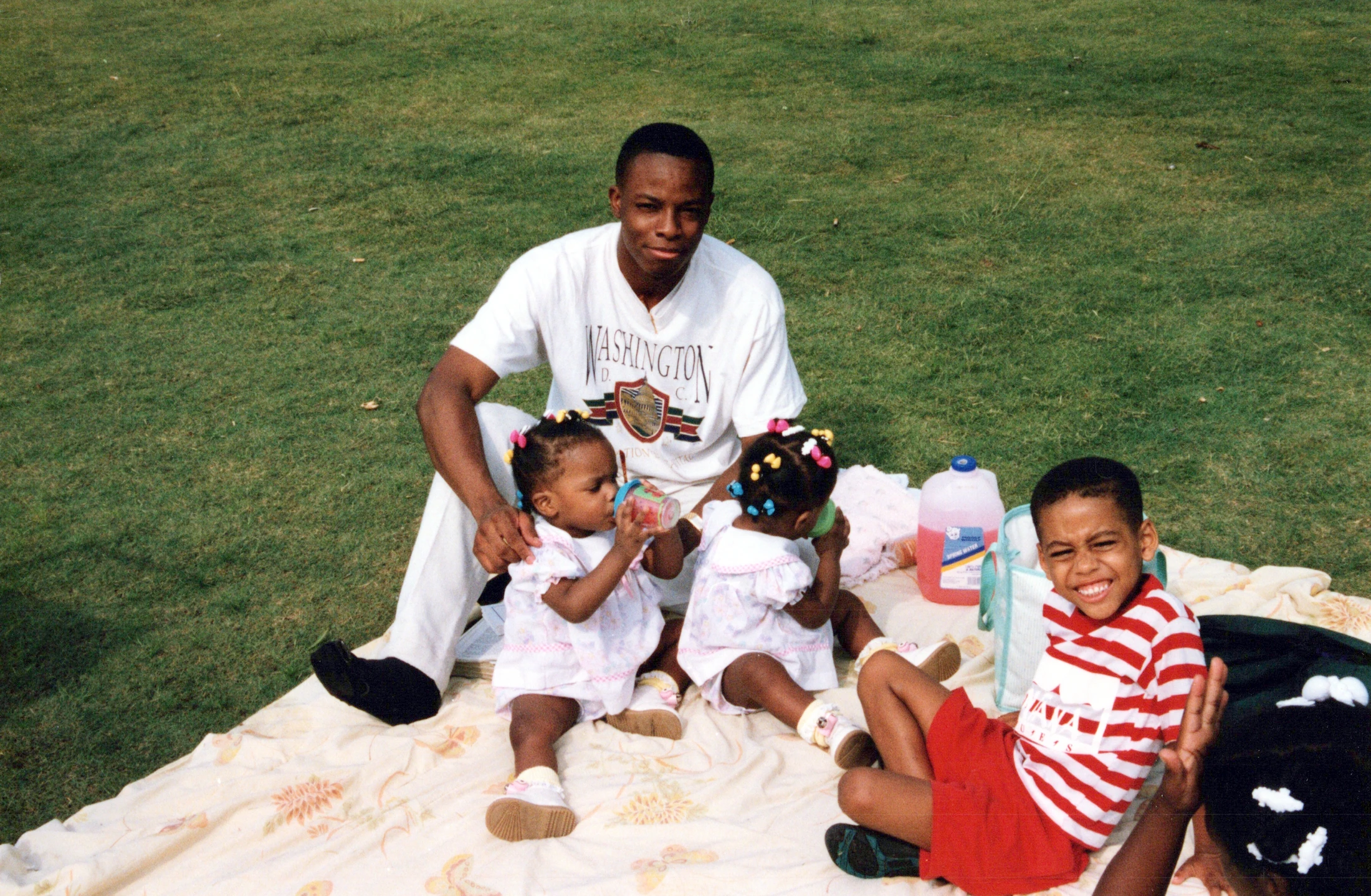A Black father sits on a blanket on the grass with his three young children, two toddlers and a young boy, enjoying a picnic on a sunny day.