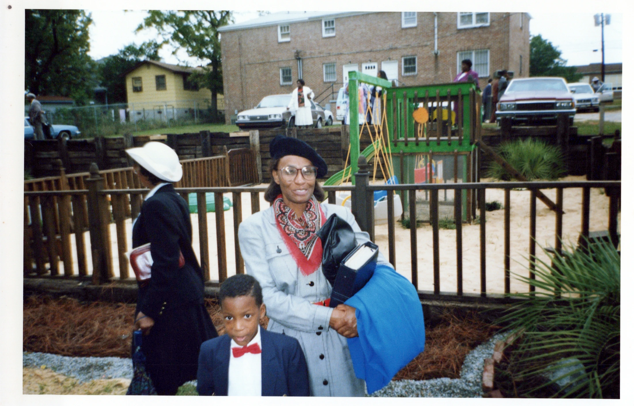 A vintage outdoor photo of a Black woman in a beret and a young Black boy in a bow tie, standing in front of a fence with a playground in the background.