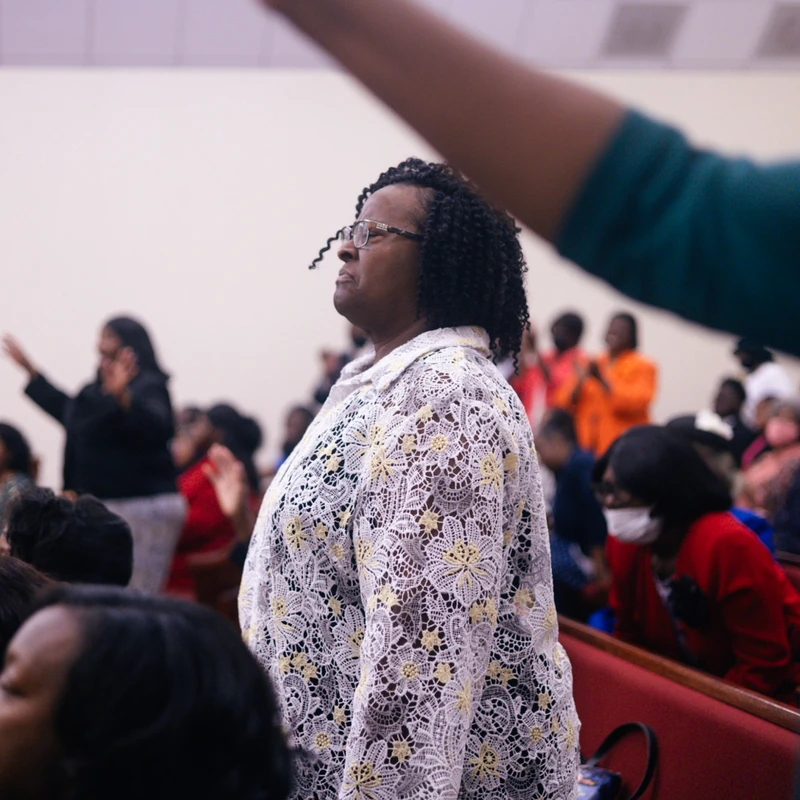 A Black woman with glasses and a white lace top stands among a congregation, looking upwards with a serene expression during a church service.