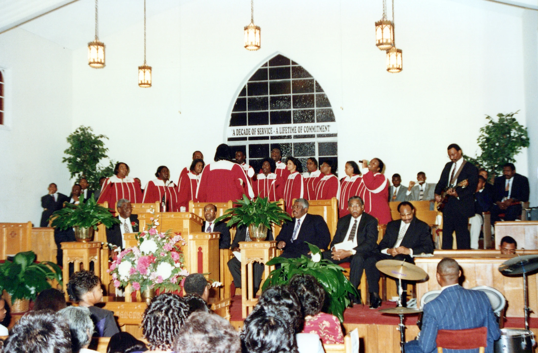 A Black church choir in red and white robes sings for a congregation. Musicians play guitar and drums on a wooden dais below a large arched window.