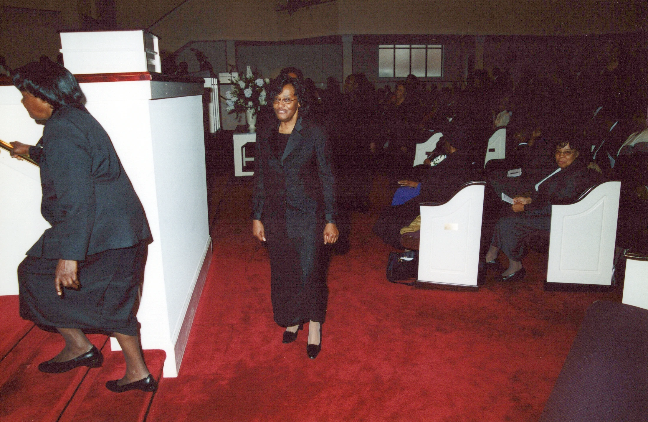 A woman in a black suit and glasses smiles as she walks down a red carpet aisle in a church. Other people are seated in white pews.