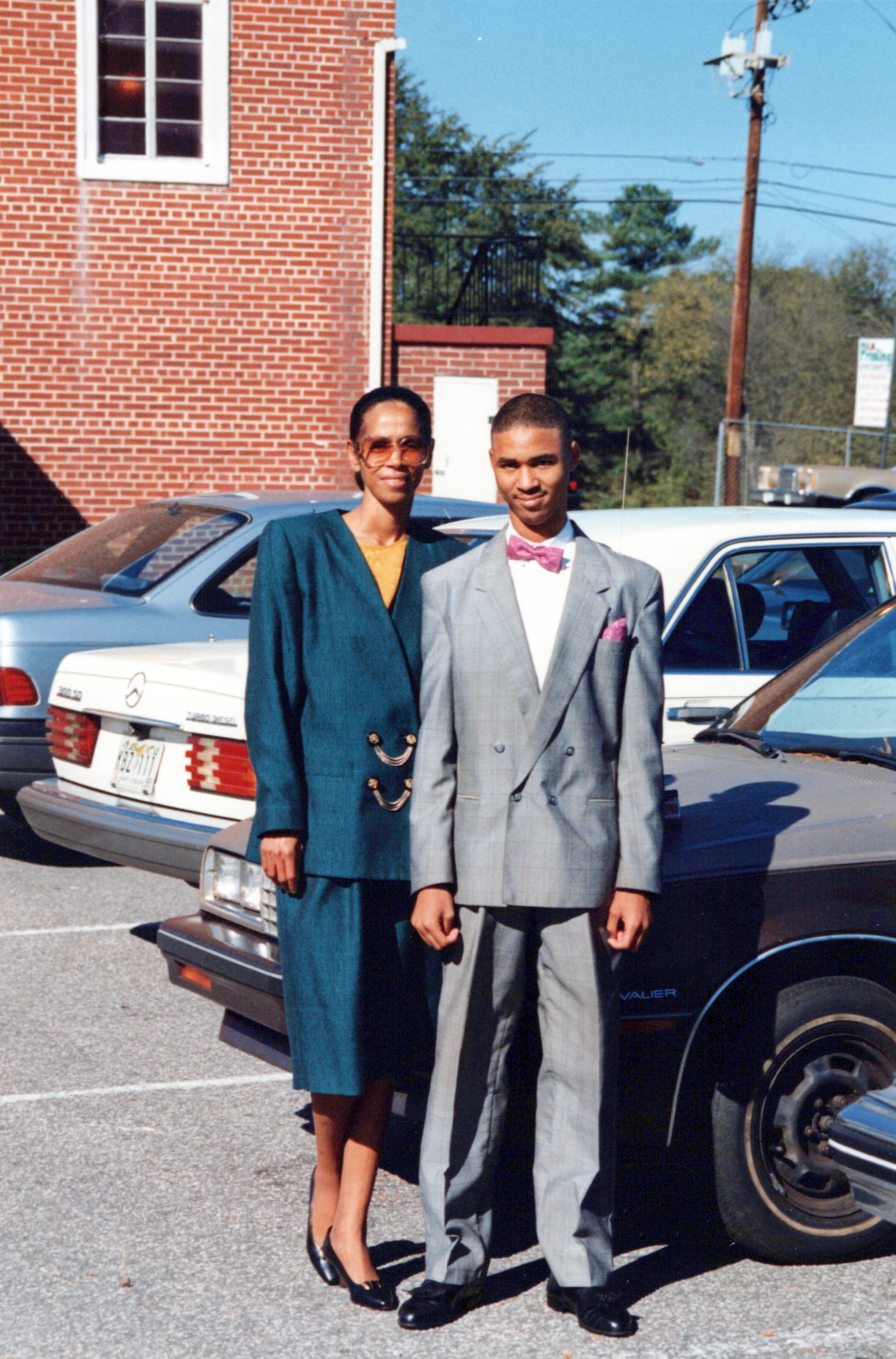A Black woman in a teal suit and a young Black man in a grey suit and pink bow tie pose for a photo in a sunny parking lot next to vintage cars.
