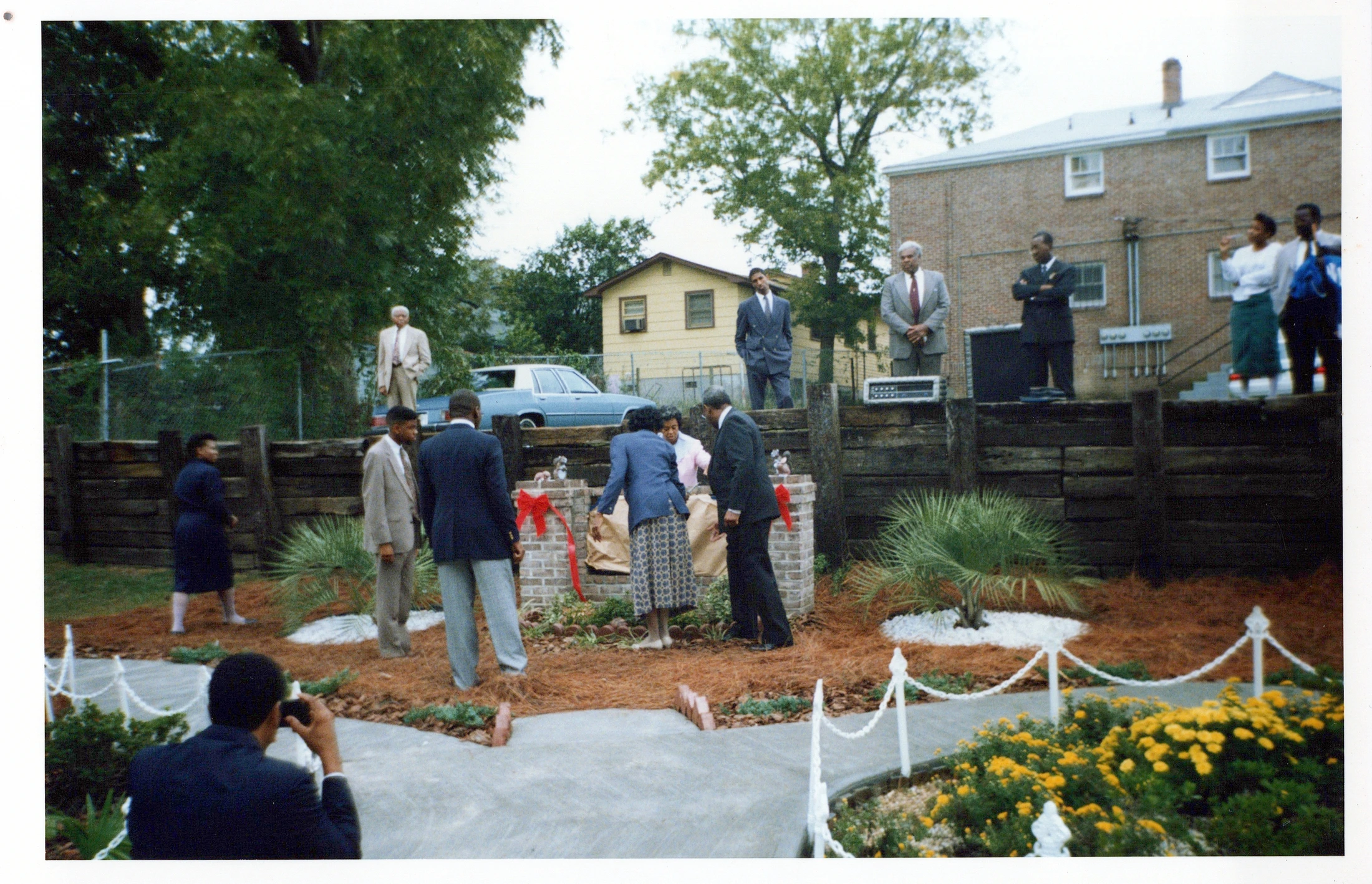 A group of African American men and women dressed in suits and dresses gather for an outdoor ribbon-cutting ceremony in a landscaped garden area.