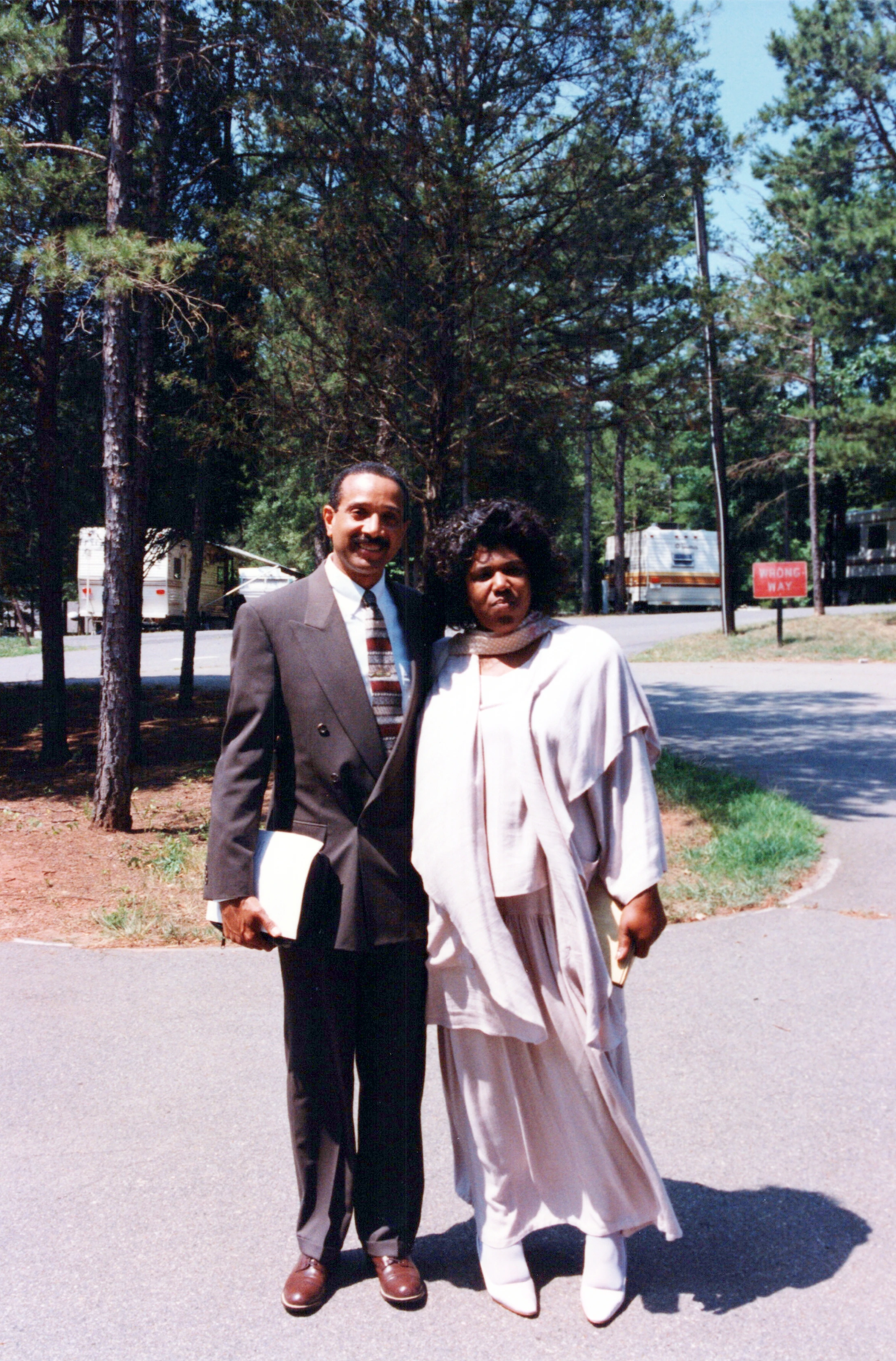 A Black man in a dark suit and a Black woman in a light-colored outfit stand together on a paved road. Behind them are tall pine trees and RVs in a park.
