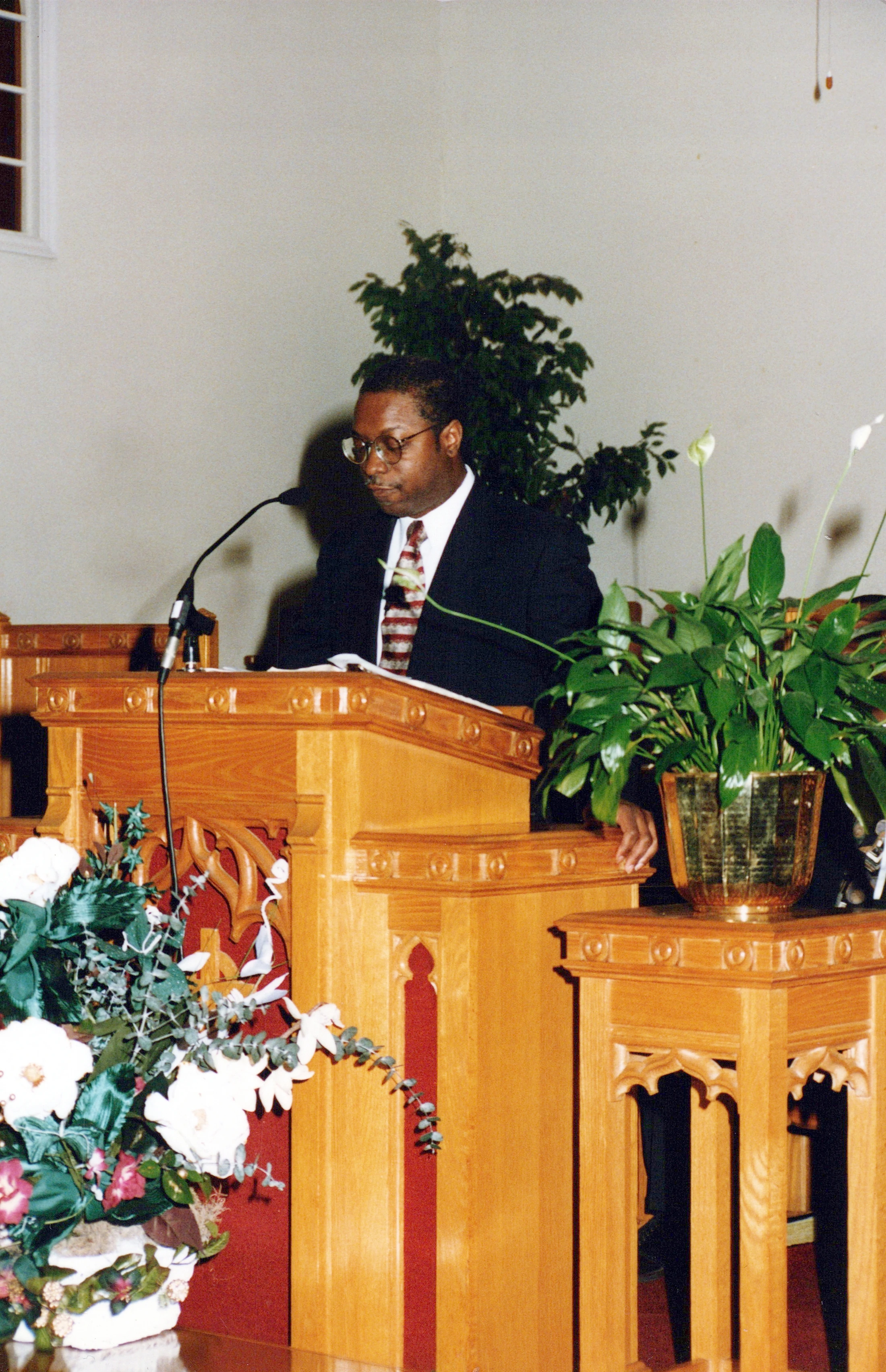 A Black man with glasses wearing a dark suit stands behind a wooden pulpit, speaking into a microphone. The pulpit is decorated with plants and flowers.
