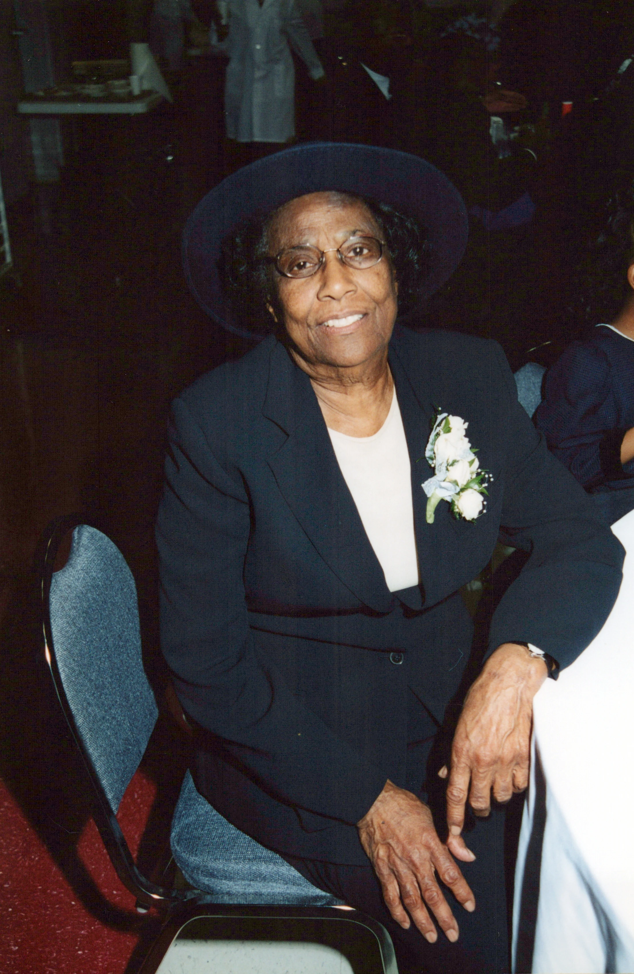 An elderly Black woman with glasses and a warm smile sits at a table. She wears a dark suit, a matching hat, and a white floral corsage on her lapel.